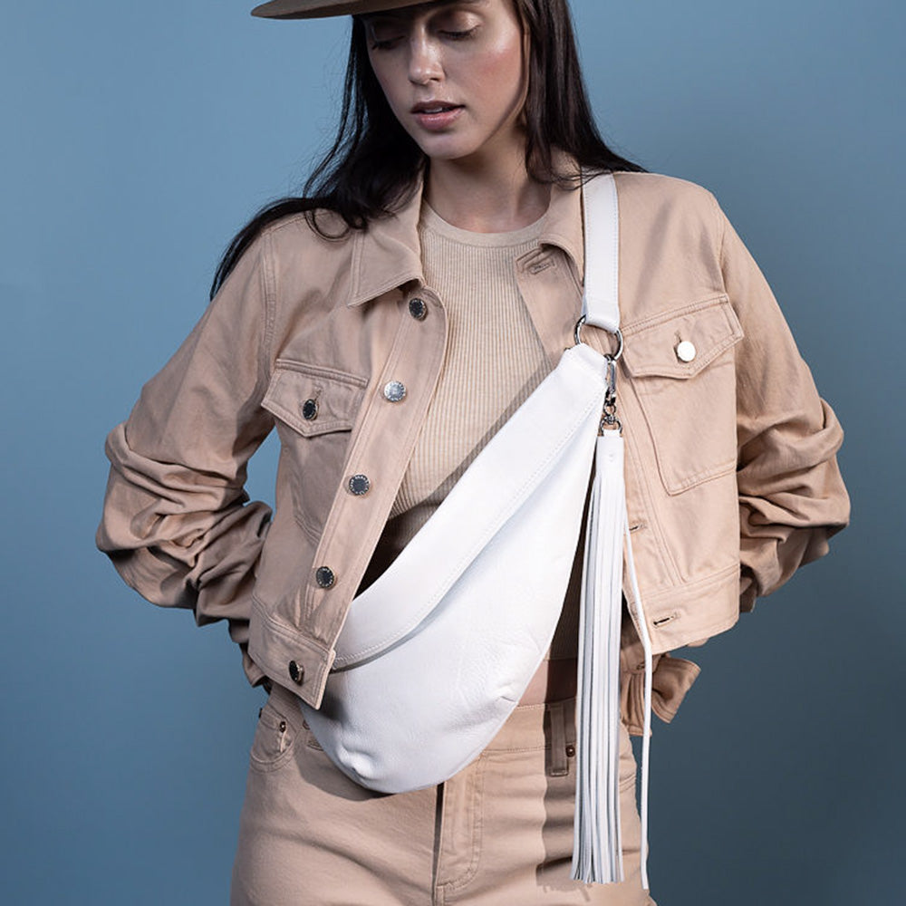 A fashionable person in a tan jacket and pants, paired with a beige top, highlights their adaptability with the Sienna - pearl white & tassel bag by evokewomen and a hat against a striking blue background.
