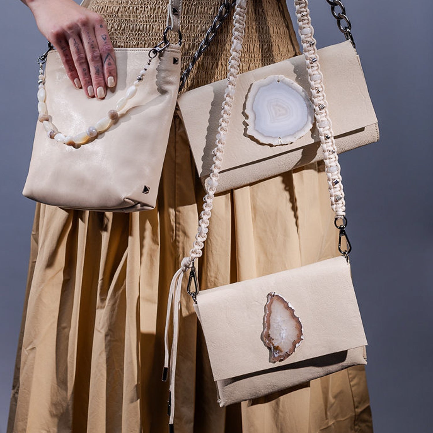 Three almond-colored BAHAMA handbags by evokewomen grace a neutral background. One features a knotted strap, another highlights an exquisite agate slice, and the third showcases chain and stone accents—all embodying timeless elegance.