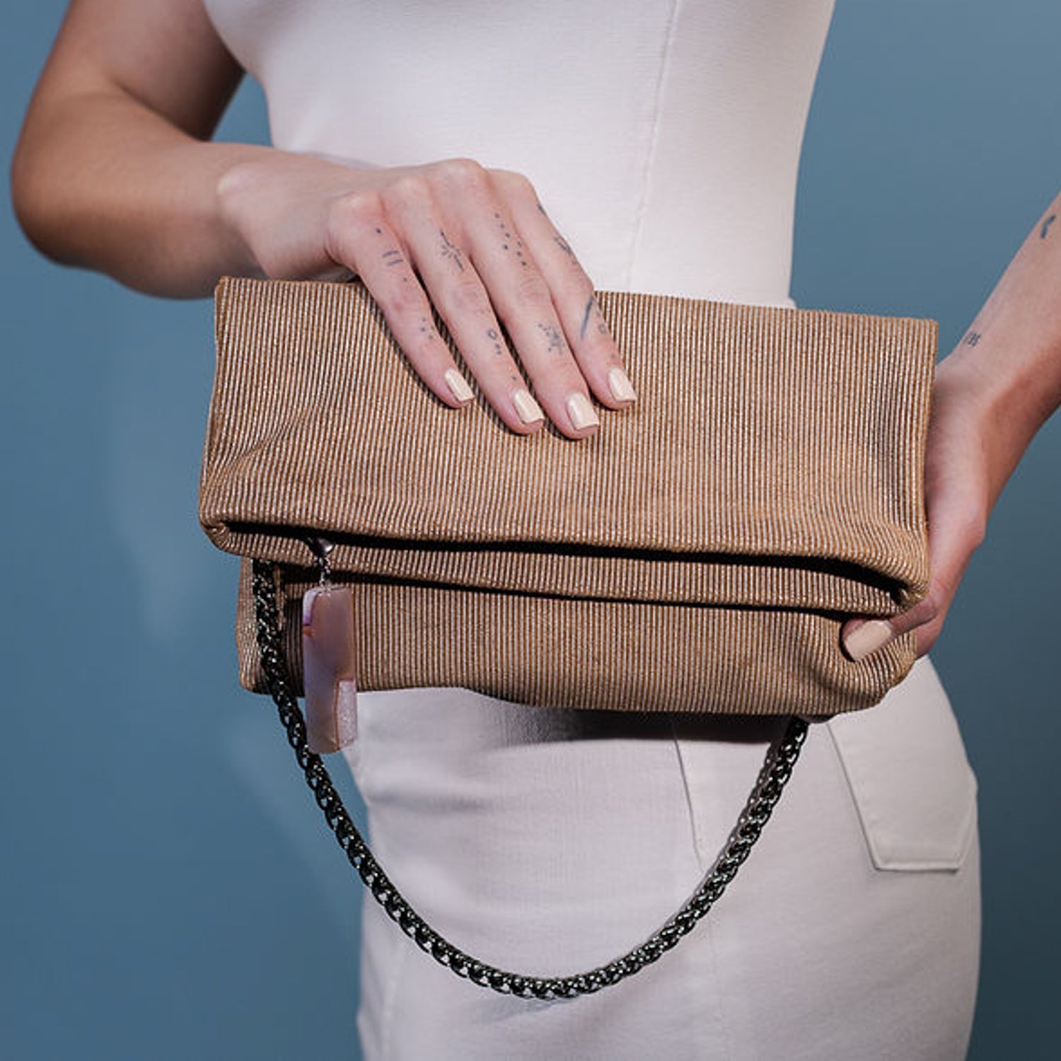 A person holds the Farah - nutshell-silver, a versatile accessory from evokewomen featuring a tan textured design with a snake spine chain strap, set against a blue background.