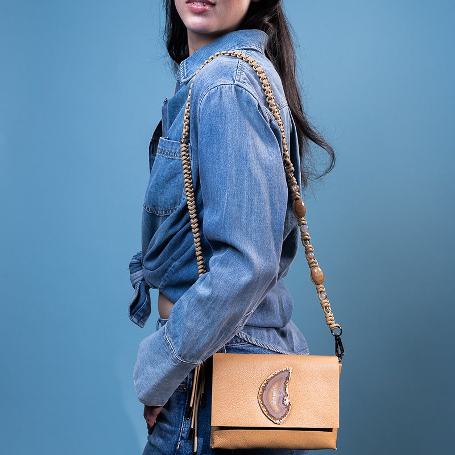 A person wearing a denim jacket and jeans holds the sophisticated BAHAMA - CAMEL purse by evokewomen, featuring a knotted strap, against a blue backdrop.