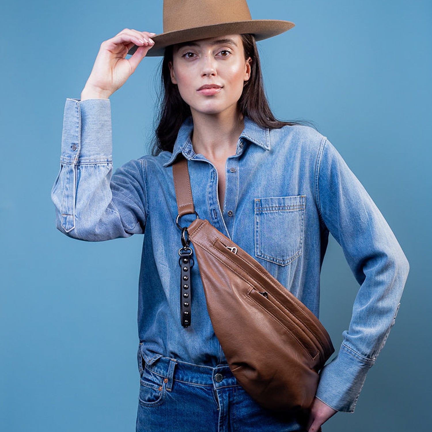 A person in a denim shirt and jeans, wearing a brown hat, holds the evokewomen Sienna - nutshell sling bag against a blue background.