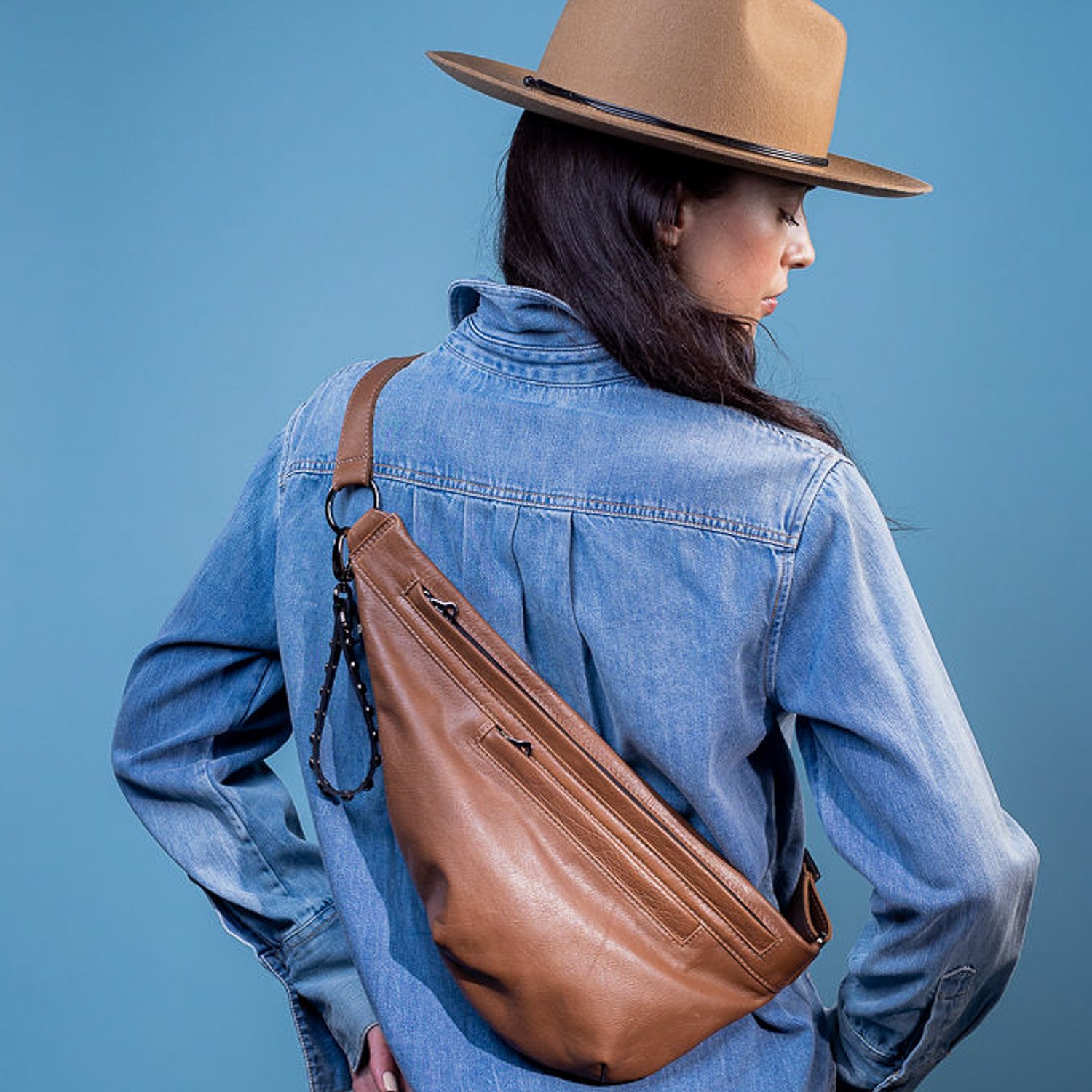 A person wearing a brown felt hat and a denim shirt carries the Sienna “nutshell” sling bag by evokewomen across their back against a blue background.