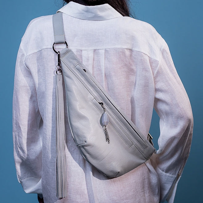 A person wearing a white shirt with an evokewomen Sienna bag in light smoke, adorned with a long tassel and metal charm, poses against a blue background.