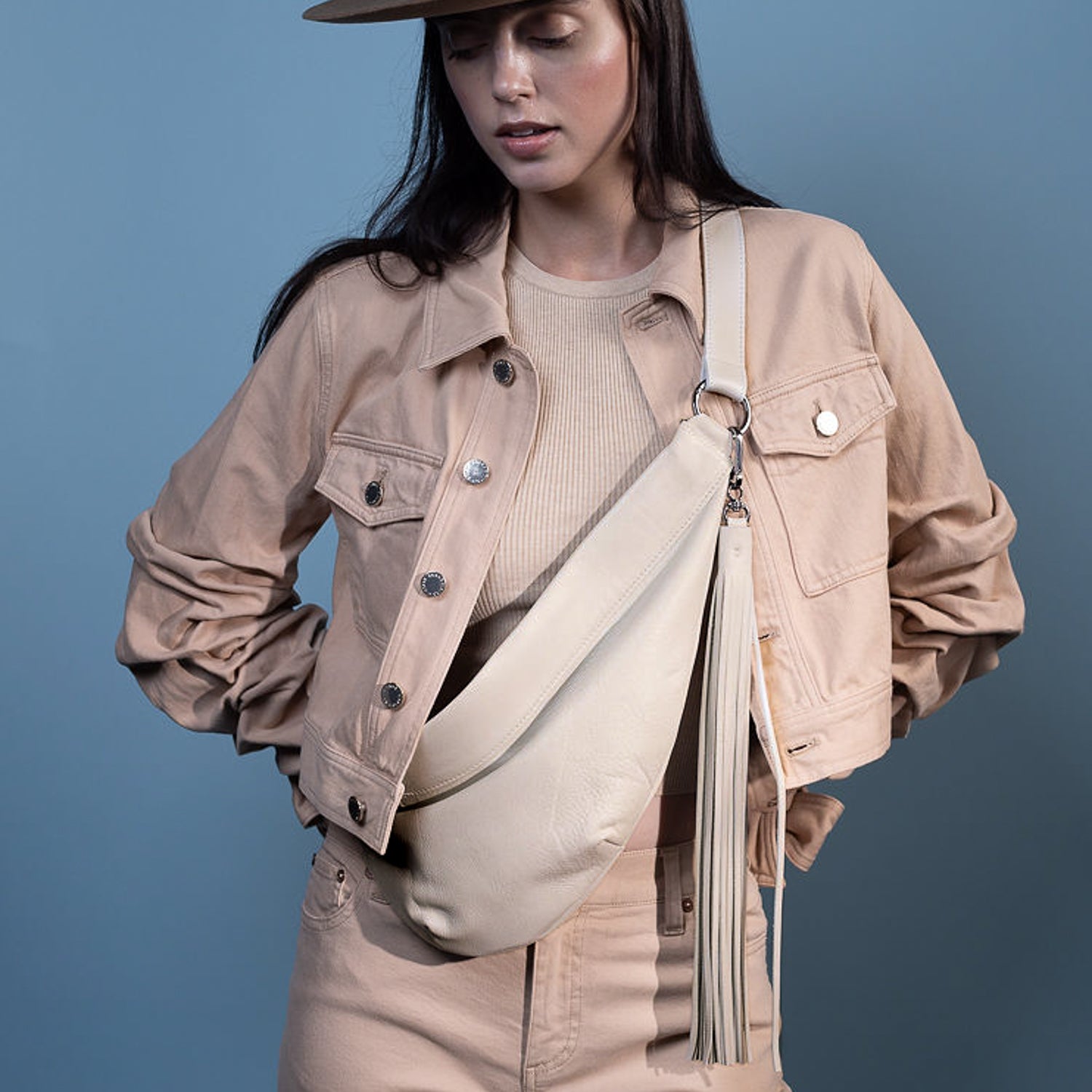 A person wearing a beige jacket and cap, stylishly accessorized with the Sienna - putty sling bag by evokewomen, stands against a blue background.