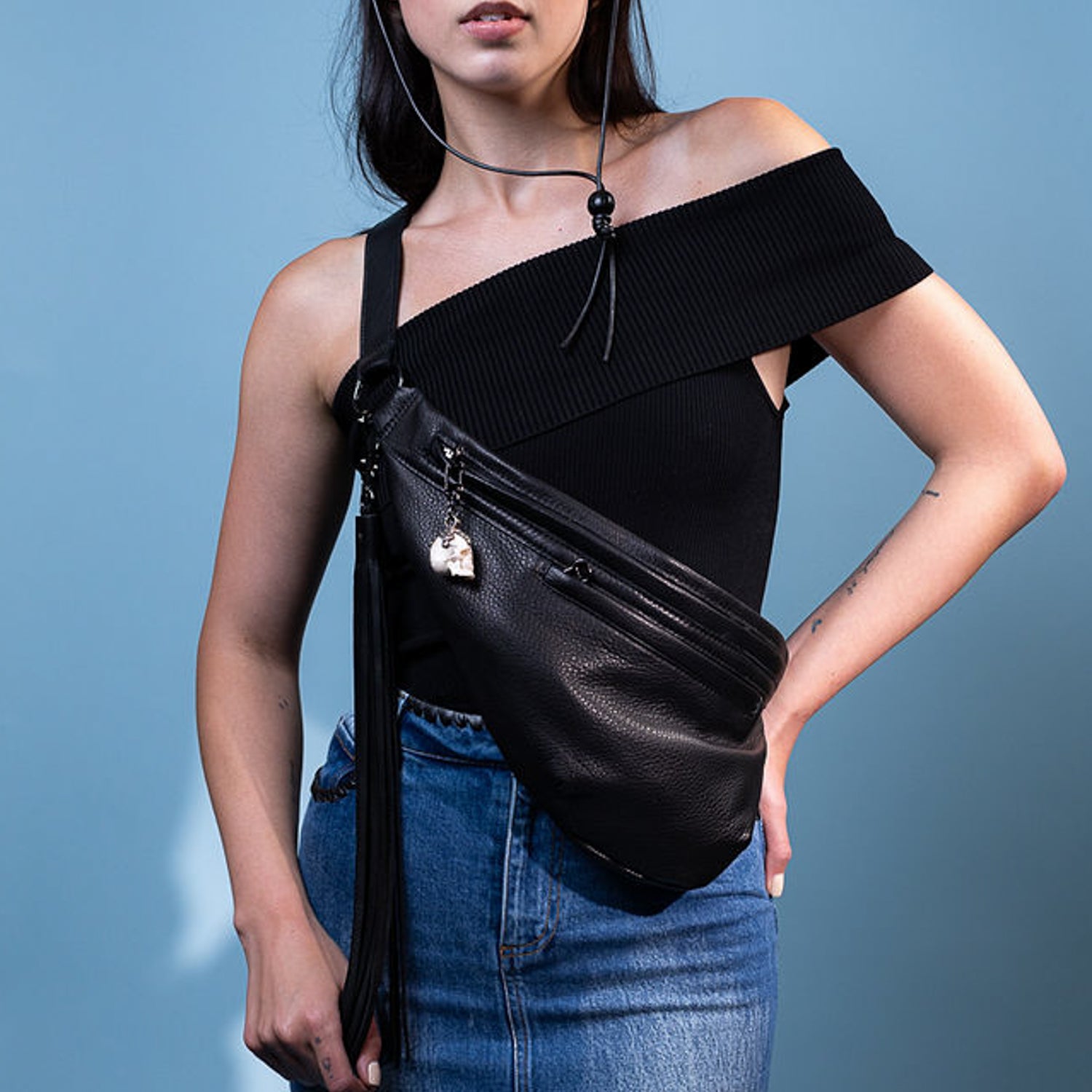 A person wearing an off-shoulder top and blue jeans, styled with the TASSEL XXL - black crossbody bag from evokewomen featuring bold gunmetal accessories, stands in front of a light blue background.