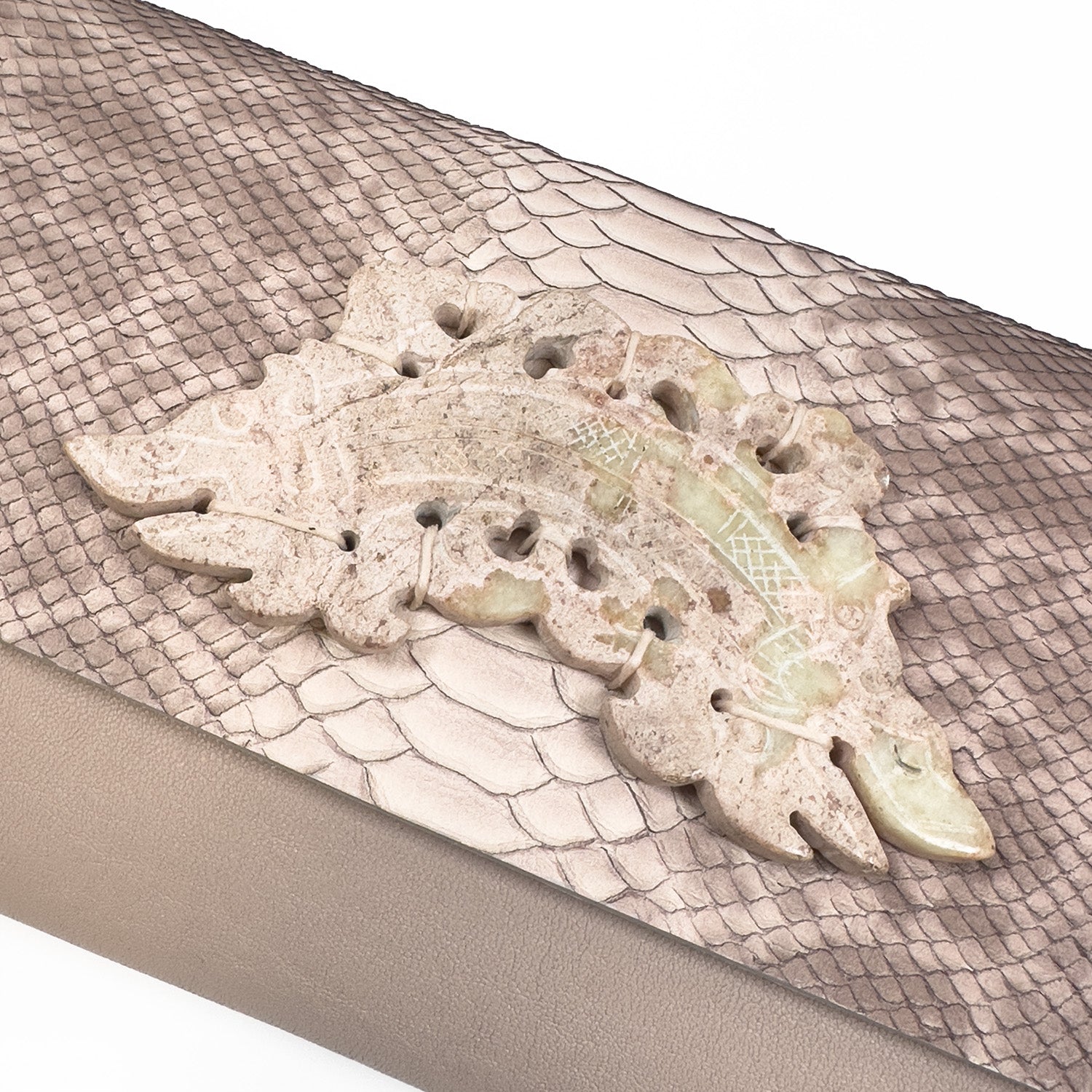 The BAGUETTE - PINK NUDE SNAKE by evokewomen, featuring intricate carvings, is elegantly placed on a textured surface that resembles an embossed snake pattern.