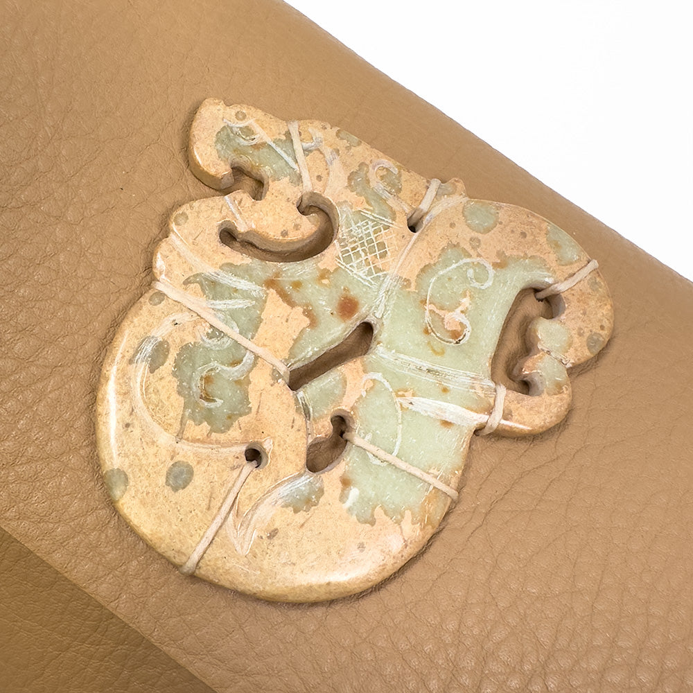 A piece from evokewomen's BAGUETTE - CAMEL collection, this vintage jade amulet features a dragon motif with beige and light green hues, set against a textured tan background. The pendant is adorned with small holes and natural patterns that reflect old-world craftsmanship.