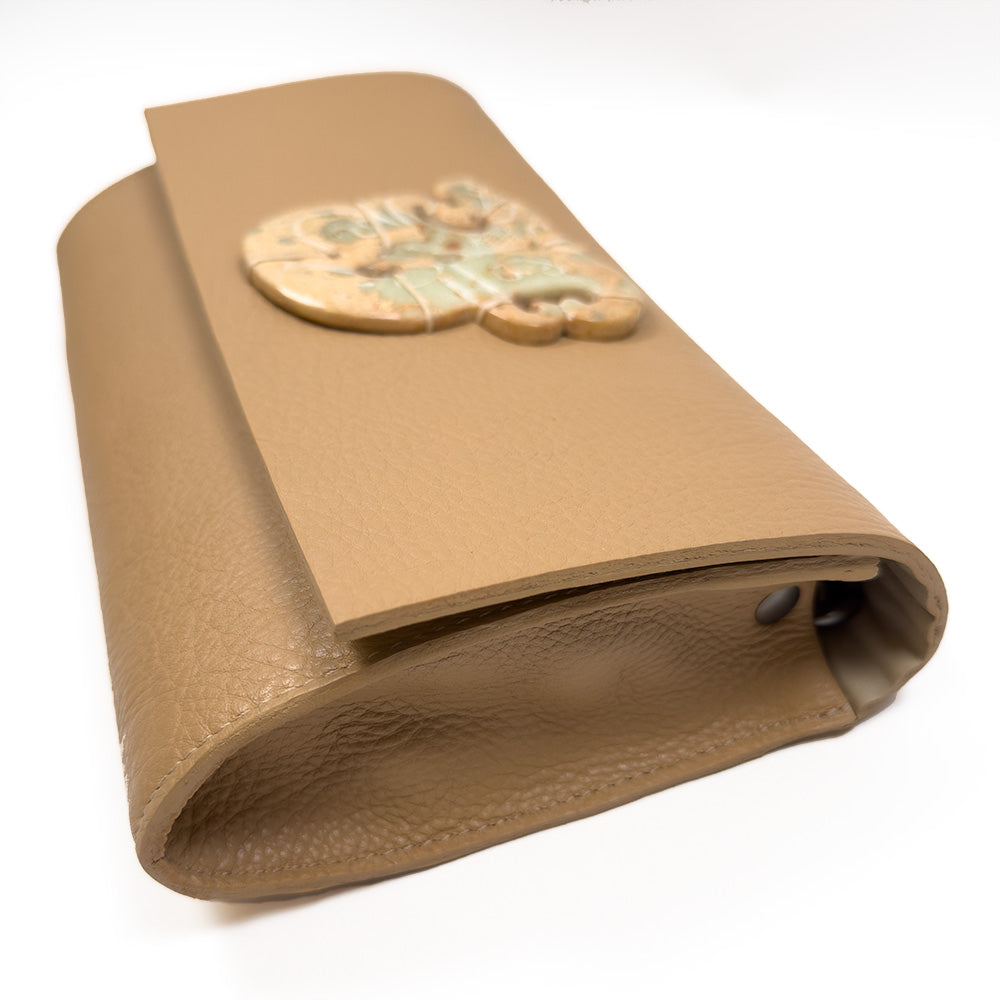 The BAGUETTE - CAMEL by evokewomen, crafted from Italian leather, features a decorative ceramic piece with a stylized face on the flap.