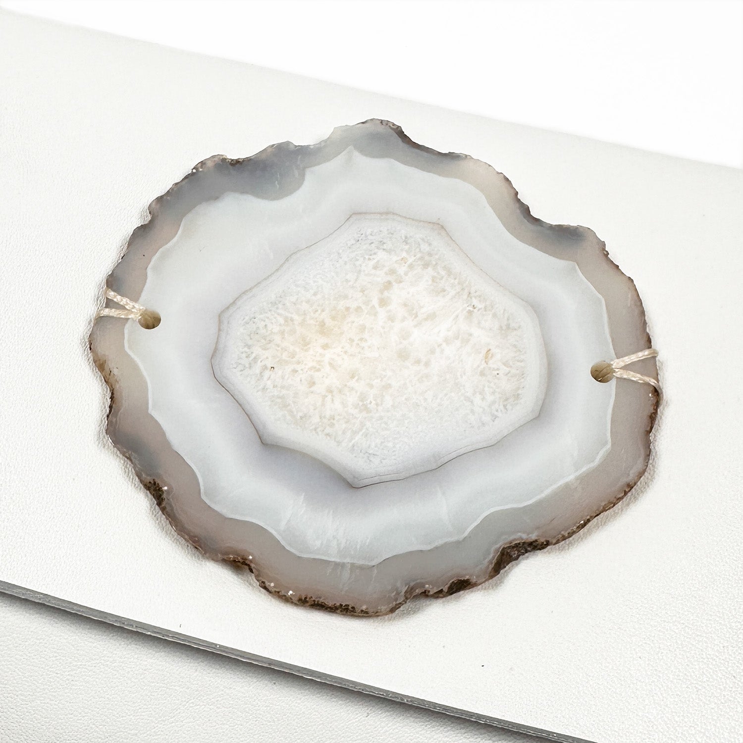 Close-up of a circular, flat-edged agate slice showcasing gray and white concentric layers, elegantly resting on a surface reminiscent of the ornate detailing found in the BAGUETTE - WHITE by evokewomen.