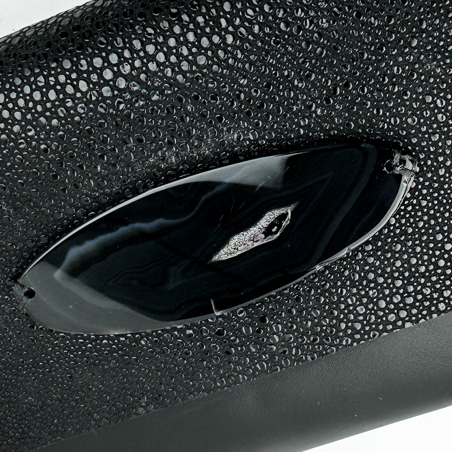 This close-up showcases the BAHAMA - BLACK STINGRAY clutch by evokewomen, crafted from luxurious Italian leather with a unique texture and adorned with a shimmering oval decoration on the front. Its exotic allure effortlessly elevates any ensemble.