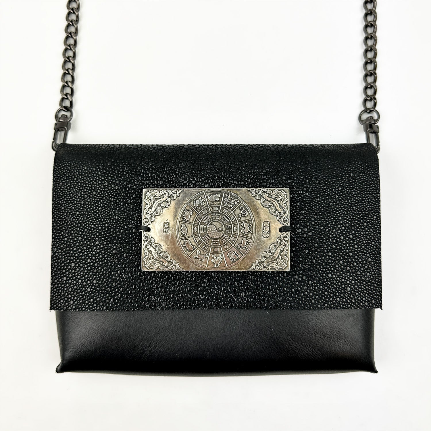 The BAHAMA - BLACK STINGRAY bag by evokewomen highlights a black textured exterior adorned with a silver rectangular decorative plate showcasing intricate circular and geometric designs. Made from Italian leather, it features a sleek crossbody chain strap that perfectly blends elegance and functionality.