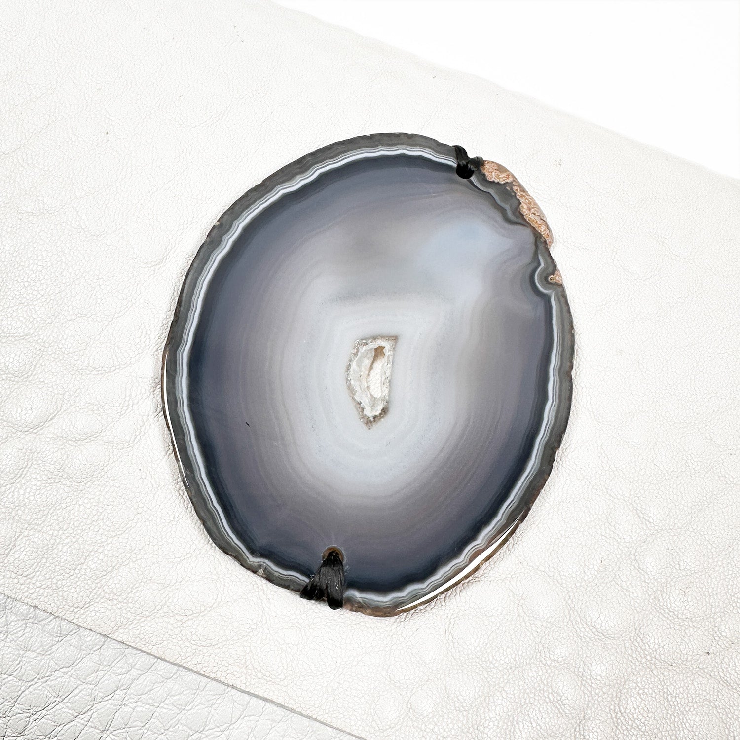 A close-up of the BAHAMA - white oval agate slice by evokewomen showcases its concentric layers in shades of gray and brown, echoing the rich tones found in Italian leather, all set against a textured white background.