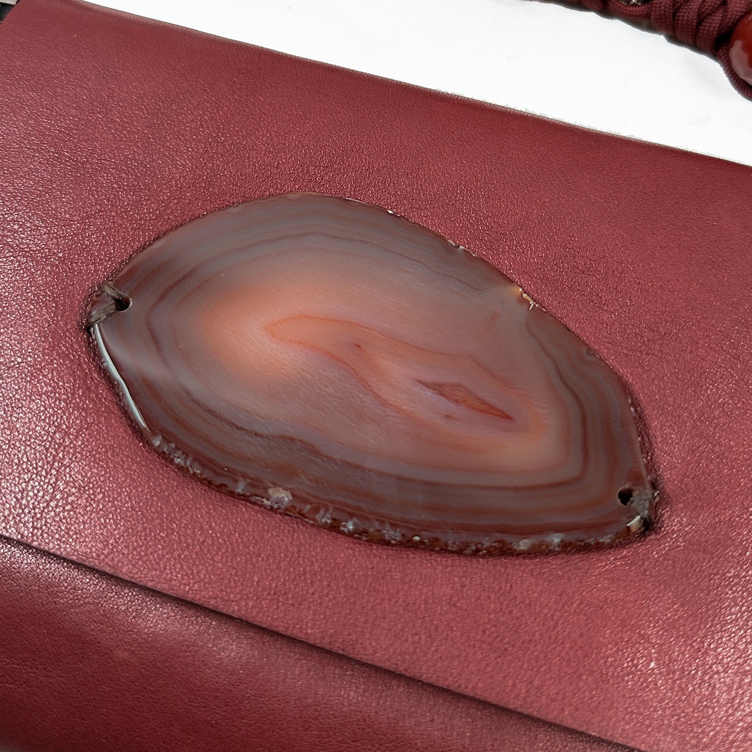 A close-up of the opulent Italian leather highlights an oval agate slice embedded within, featuring concentric bands of reddish-brown tones, echoing the sophisticated elegance embodied by the evokewomen BAHAMA - wine bag.