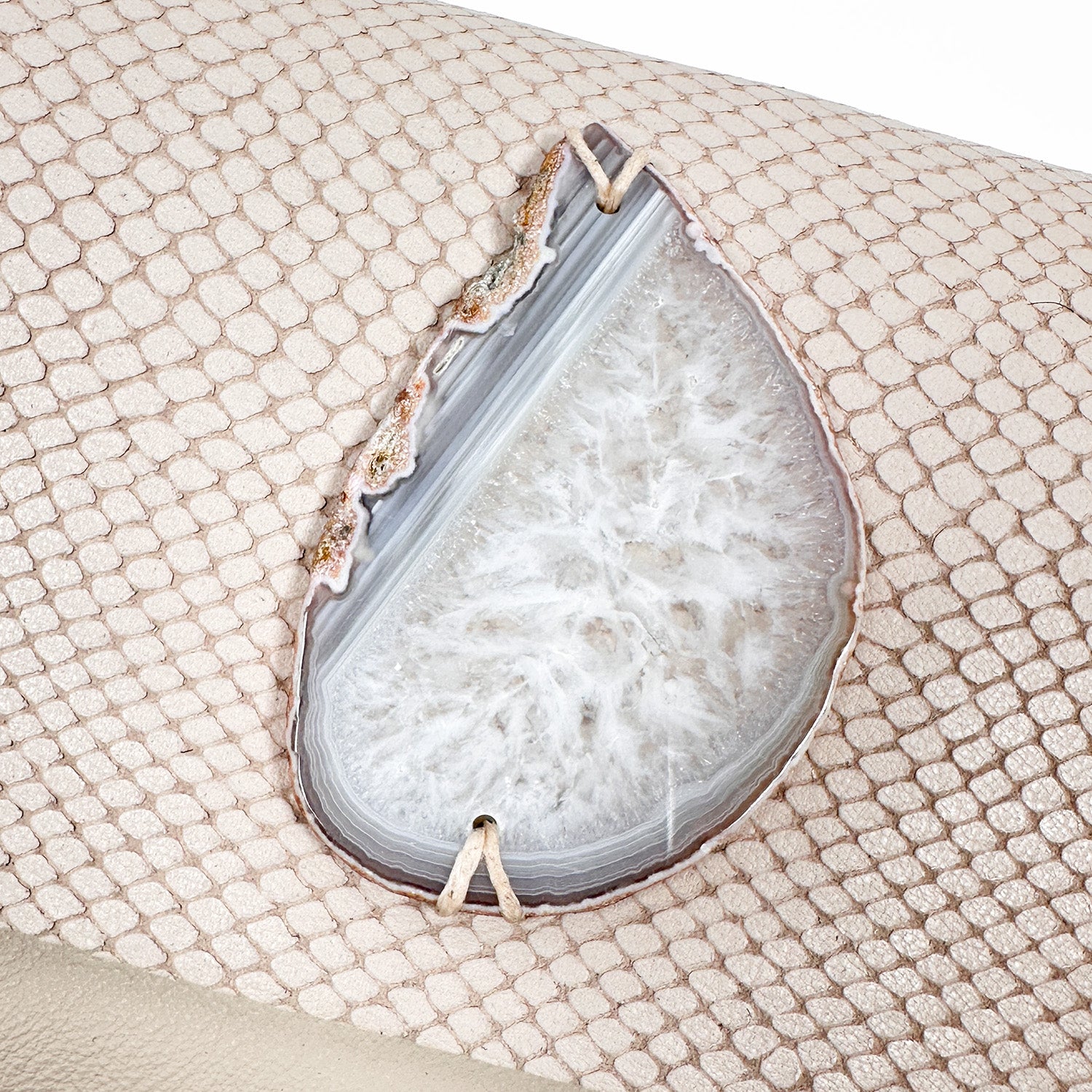 Close-up of a teardrop-shaped white druzy agate stone set into a powder beige snakeskin-embossed leather surface, showing intricate crystal formations and delicate prong detailing.