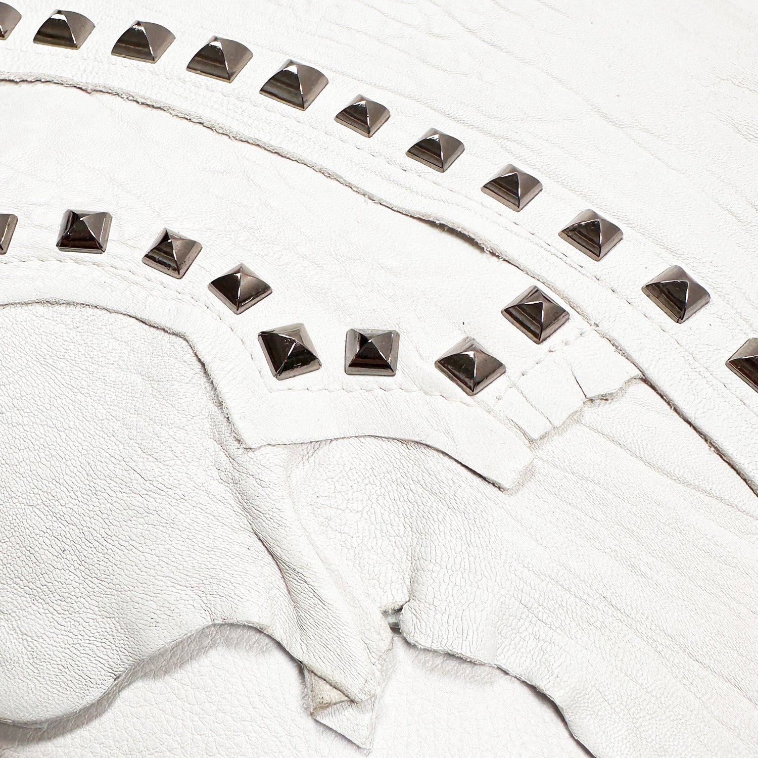 A close-up of the DRITA - white crossbody bag by evokewomen showcases luxurious Italian leather embellished with decorative silver studs in elegant curved patterns, complete with an adjustable strap.