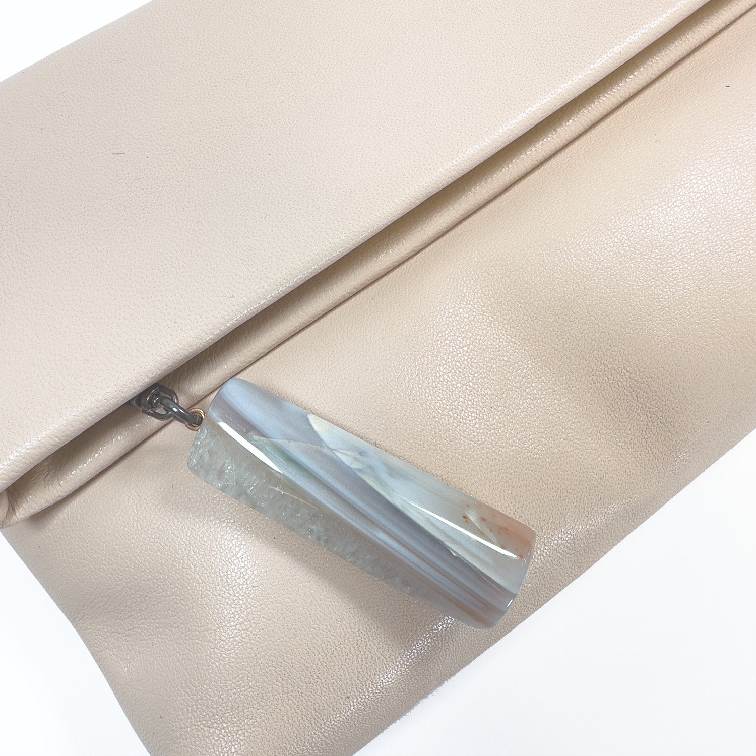 The Farah clutch by evokewomen in almond boasts a striking rectangular stone pendant and a delicate snake spine chain, adding an edgy flair.