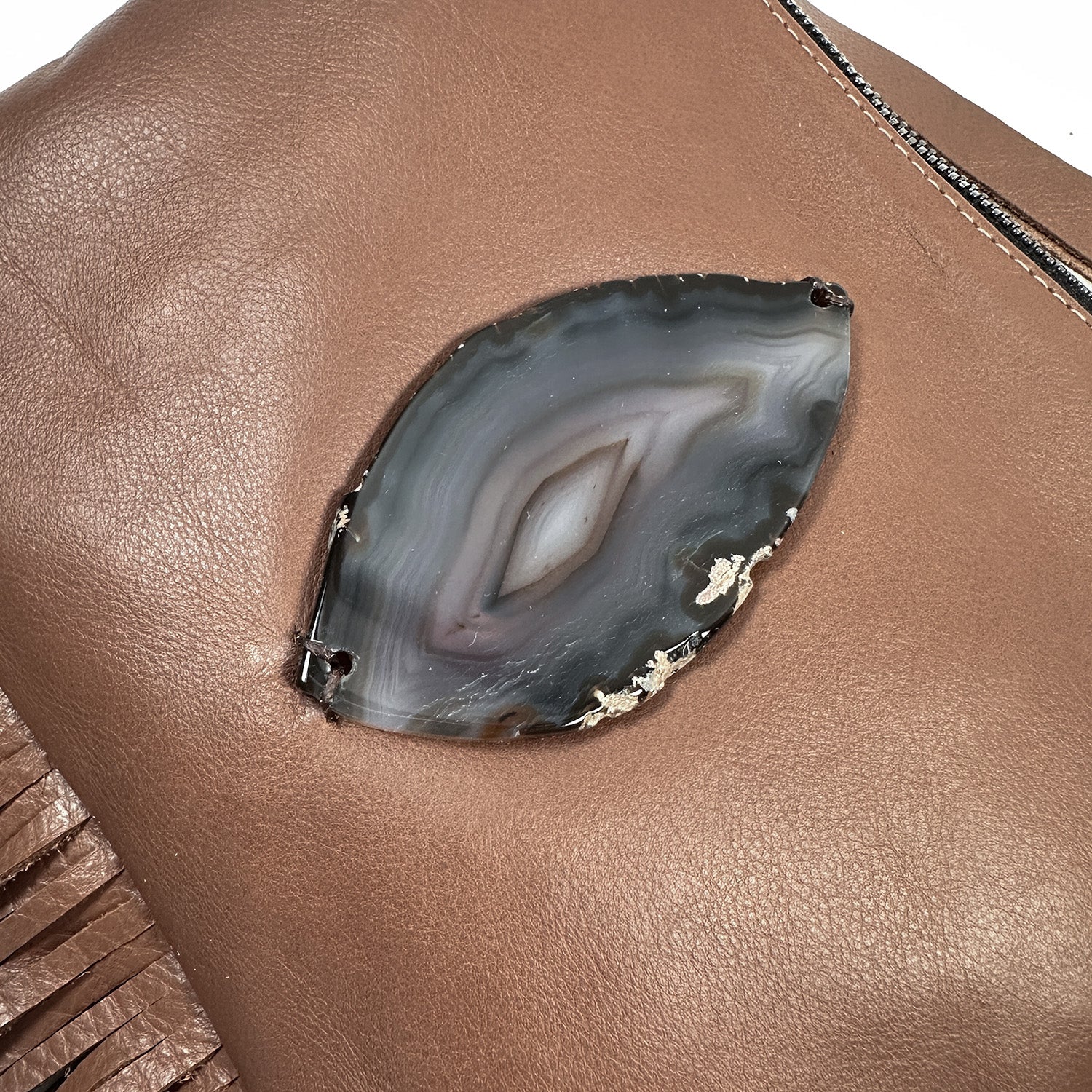 Close-up of the evokewomen Fringe bag - nutshell, adorned with brown leather fringes and featuring a decorative stone with an eye-like pattern set into the material.