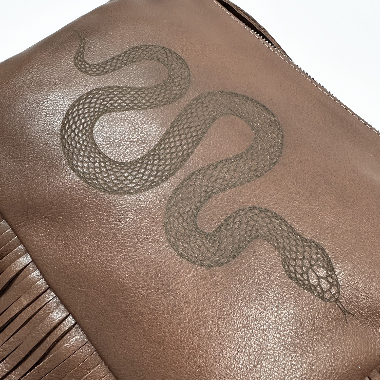 The Fringe bag - nutshell laser snake by evokewomen showcases a brown leather festival design with a snake pattern and leather fringes on the left side.