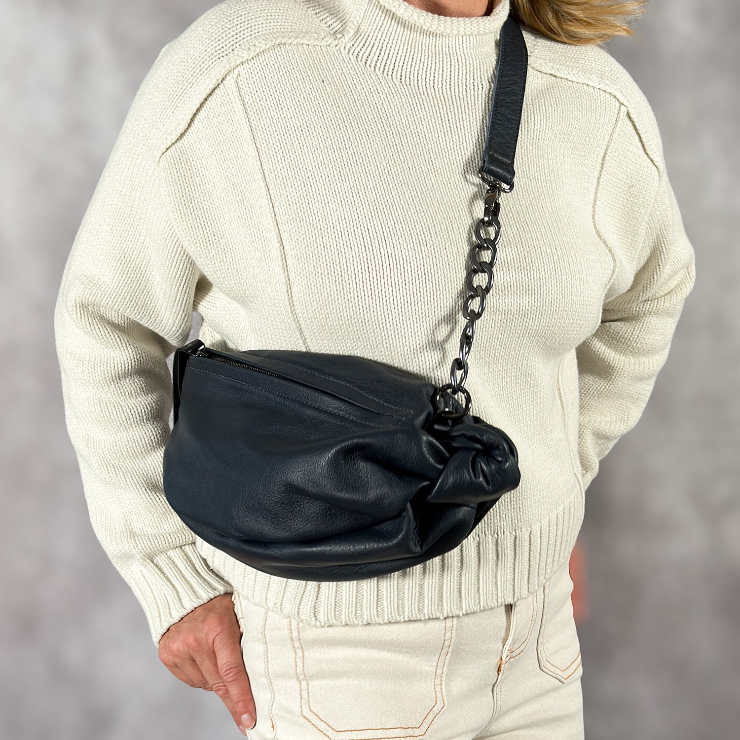 A person wearing a cream sweater and light pants holds an evokewomen LONDON - BLACK, a luxury leather bag featuring a chic chain strap.