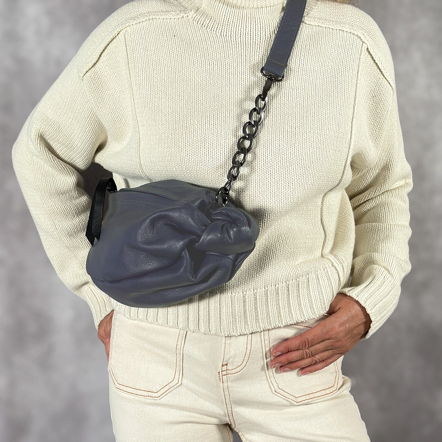 A person in a white sweater and beige pants holds the chic LONDON - NAVY cross body bag by evokewomen, featuring a gray leather chain-strap, against a gray backdrop.