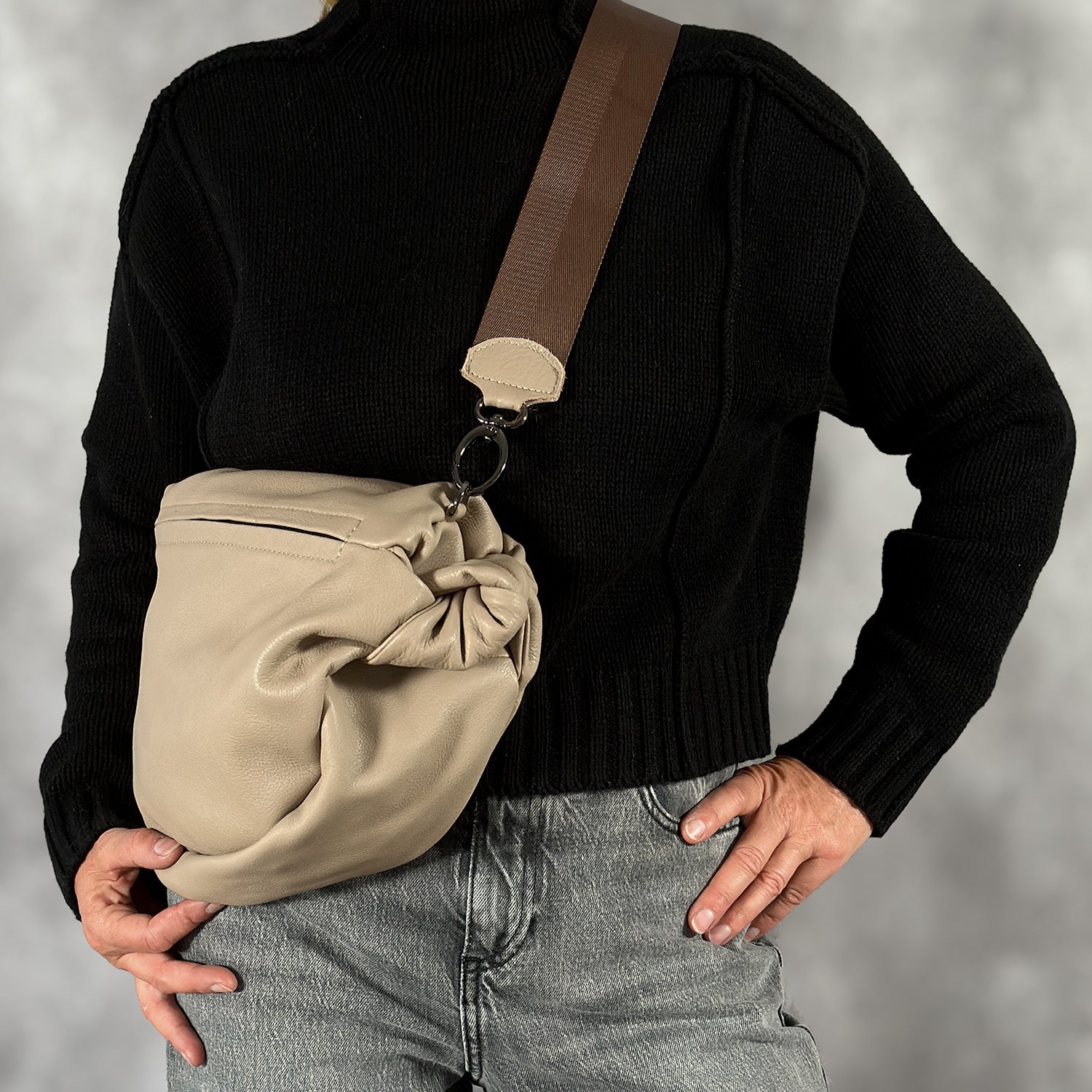 A person in a black sweater and gray jeans holds the LULA - SAND slouchy sling bag from evokewomen, featuring a wide brown strap.