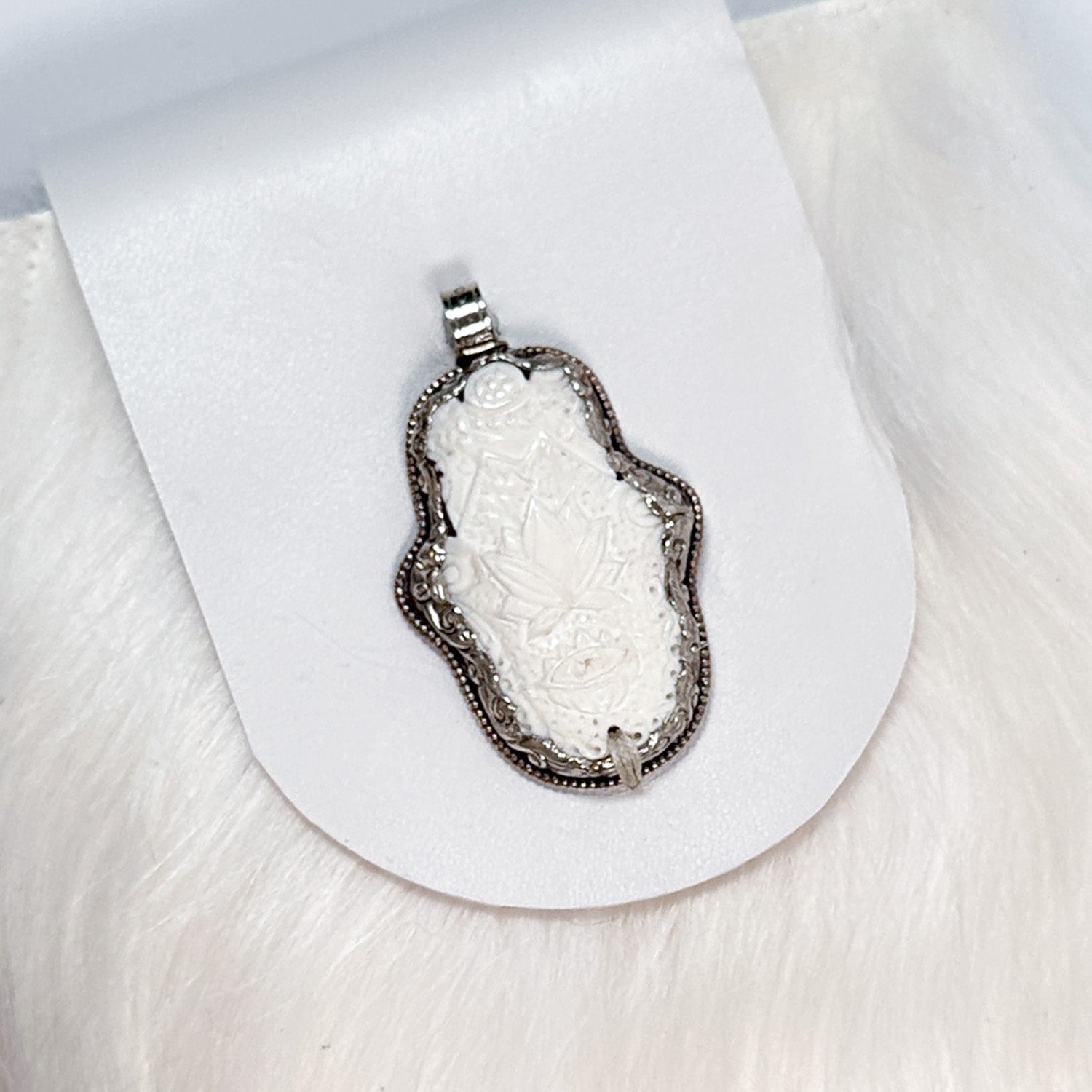 The "MIA - WHITE" pendant by evokewomen features an intricately carved floral design, reminiscent of a Tibetan bone amulet, beautifully bordered in metal and showcased on a white background.