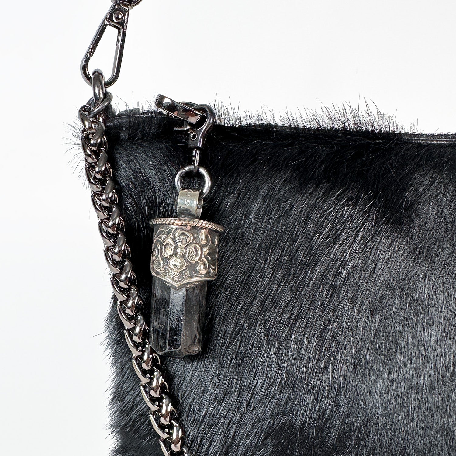 Close-up of the luxurious POPPY - BLACK shoulder bag by evokewomen, featuring a goat fur texture with a silver chain strap and adorned with a decorative pendant showcasing a dark crystal encased in patterned silver casing.
