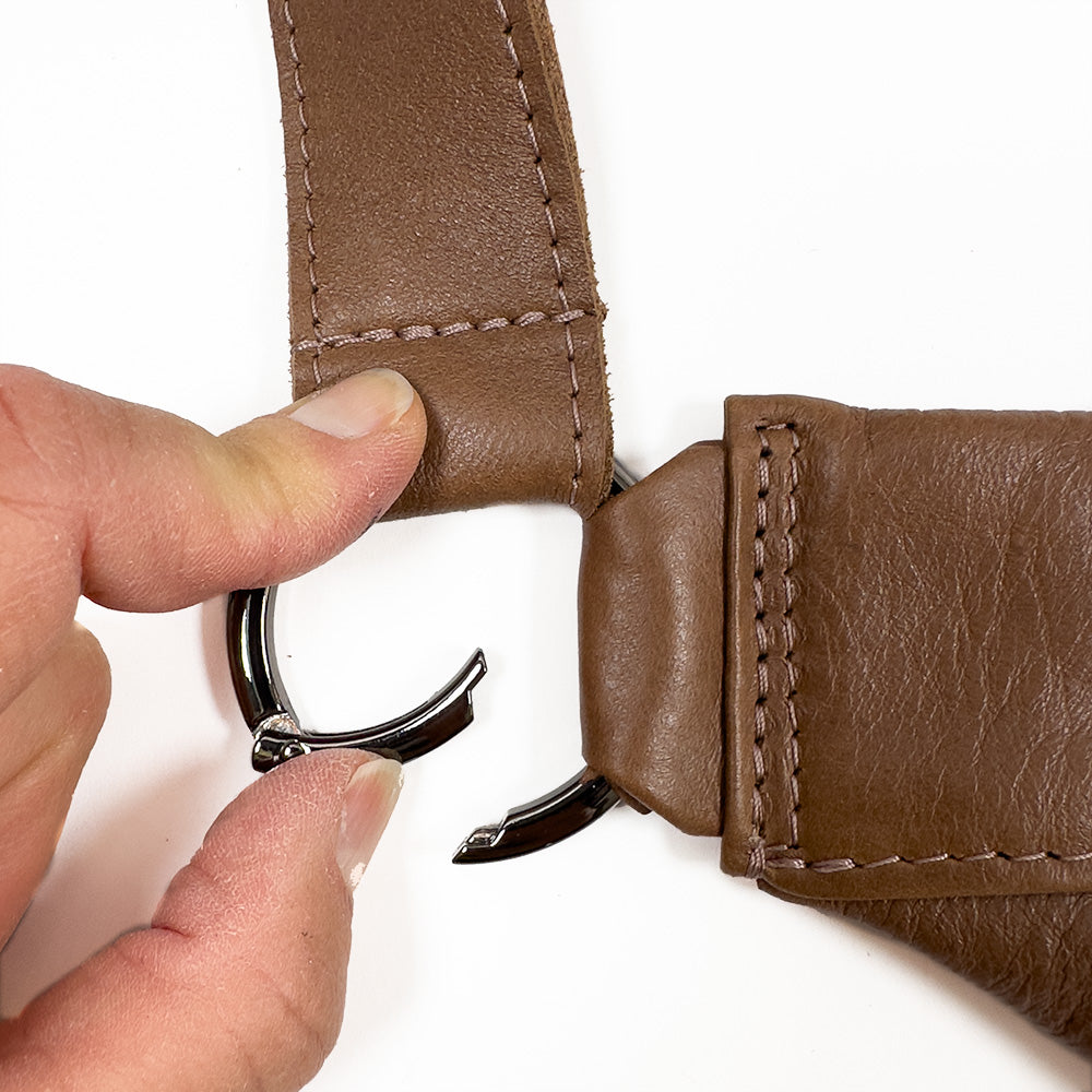 Close-up of a hand fastening a metal clasp to the brown leather strap of an evokewomen Sienna - nutshell bag.