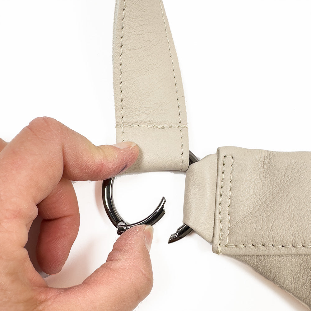 A hand clasps a silver metal ring attached to the beige leather strap of an elegant evokewomen Sienna - putty bag.