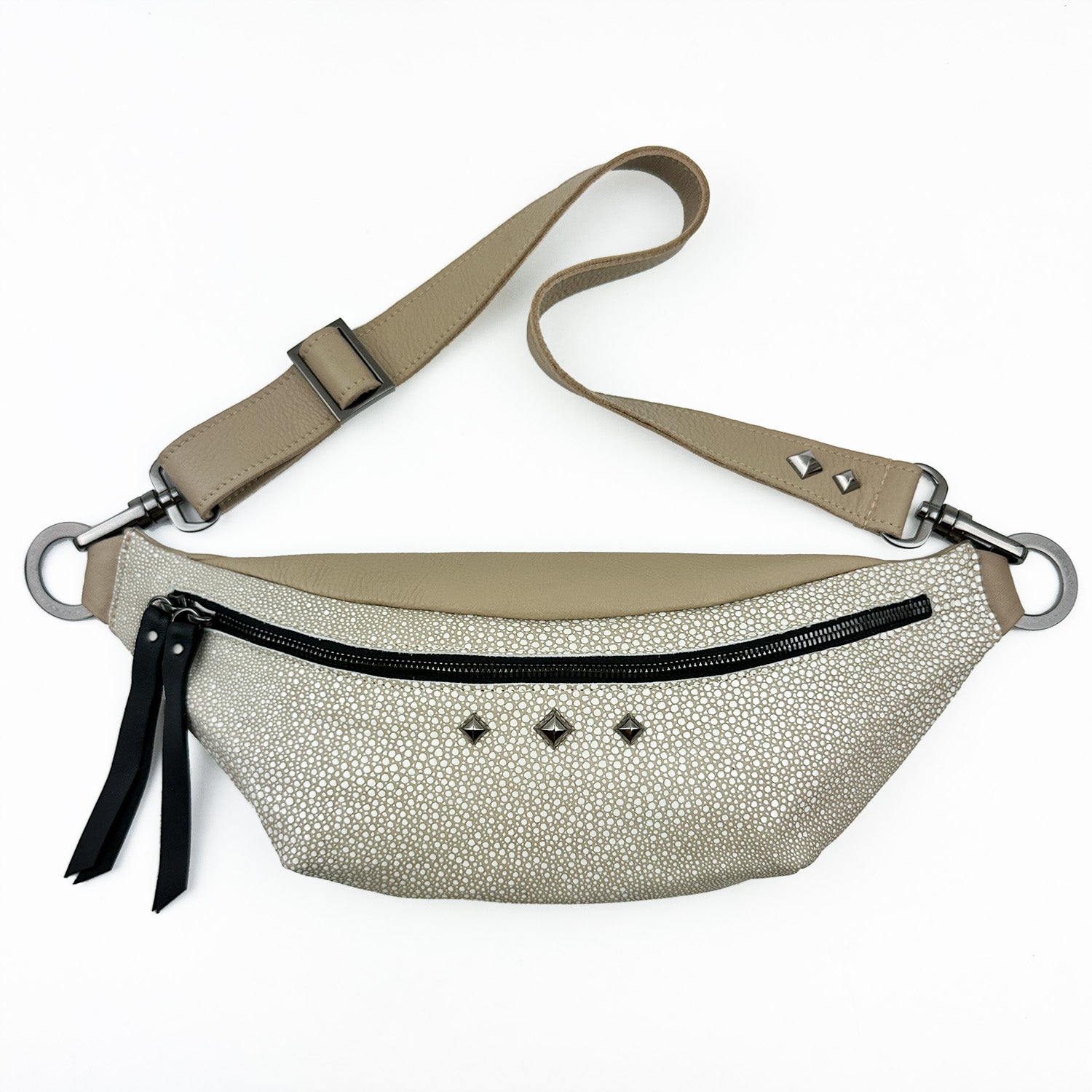 Presenting the STELLA - SAND STINGRAY by evokewomen: a stylish waist bag in beige and white with a textured finish. It features a black zipper, chic black tassels, and subtle small square studs. The adjustable strap with metal rings allows you to wear it cross-body for a versatile look.