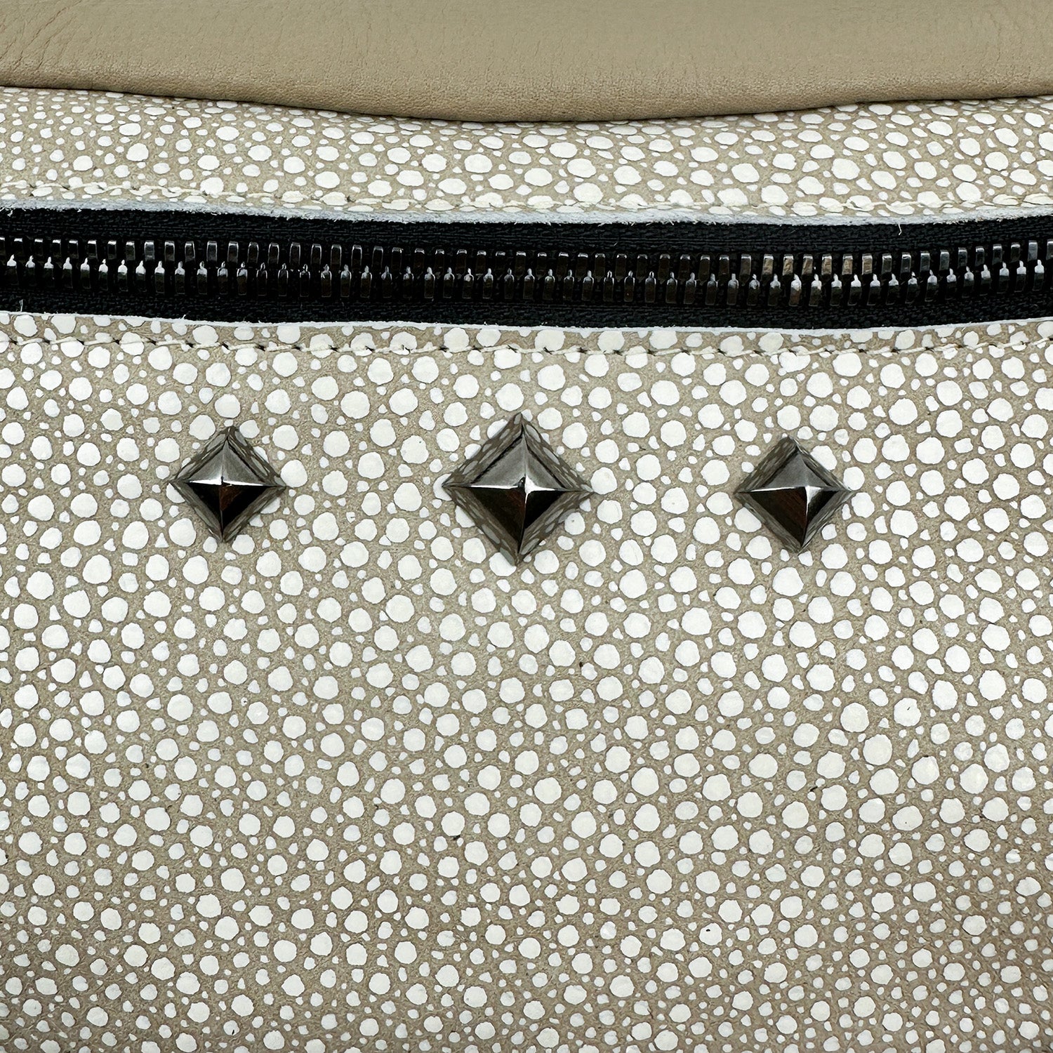 A close-up of the sand stingray-textured leather surface on the sleek STELLA crossbody bag by evokewomen, showcasing three metallic pyramid studs and a black zipper.