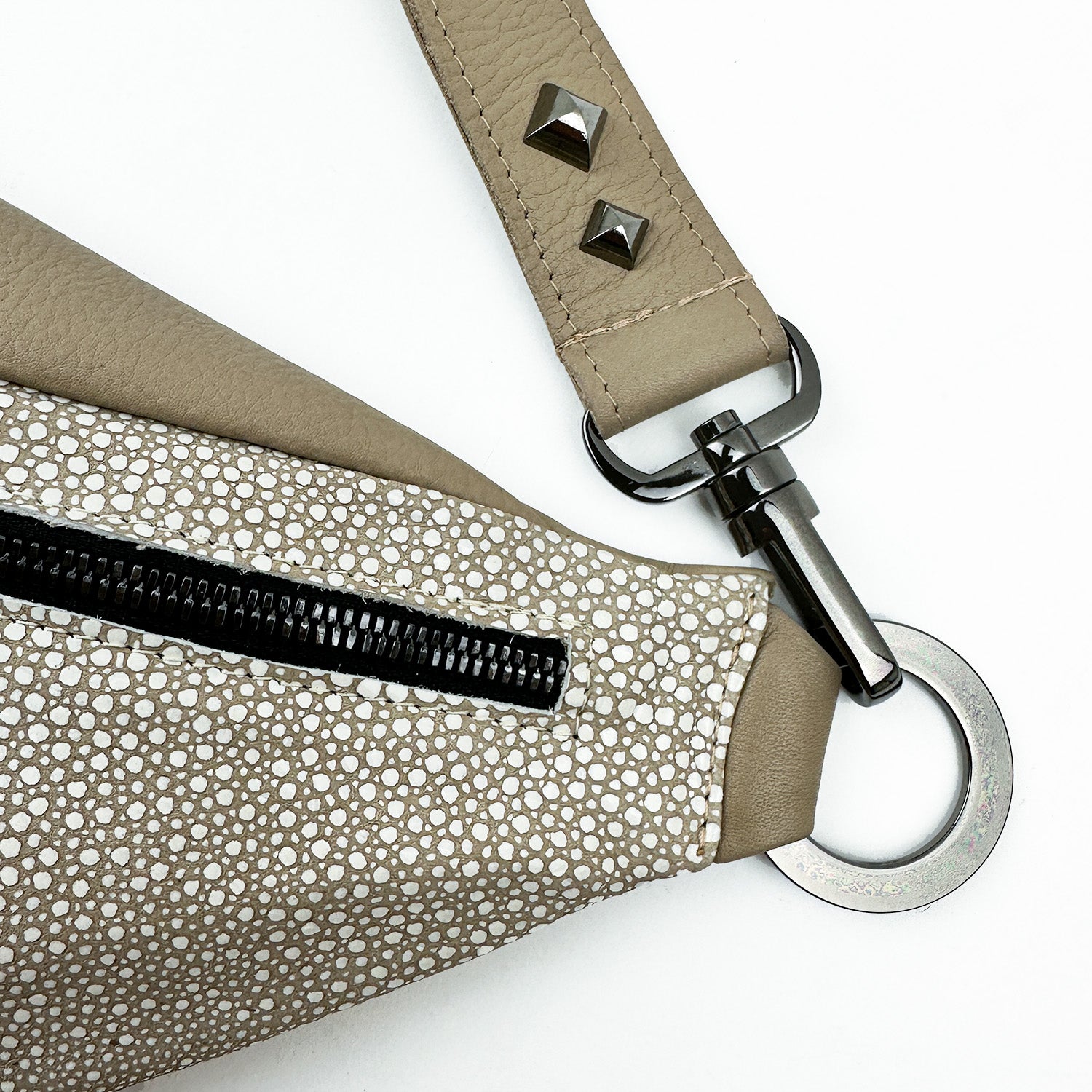 Close-up of the STELLA - SAND STINGRAY cross body bag by evokewomen, showcasing its beige strap adorned with metal studs and a circular ring attachment, featuring a textured pattern and a black zipper.