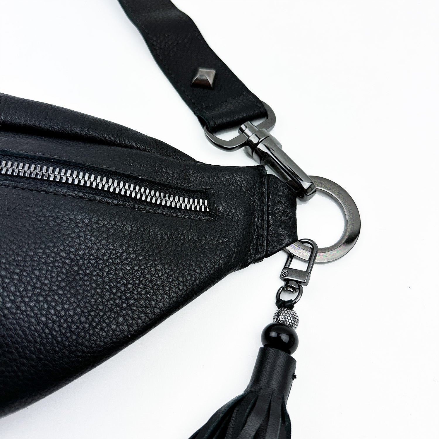 Close-up of the stylish STELLA - BLACK sling bag by evokewomen, crafted from sleek black leather and featuring a zippered pocket, metal ring and clip, and an elegant tassel detail.