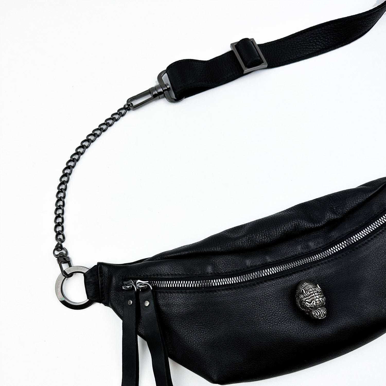 The STELLA - BLACK by evokewomen is a black leather waist bag with an adjustable strap and metal chain, adorned with a decorative skull emblem and zipper.