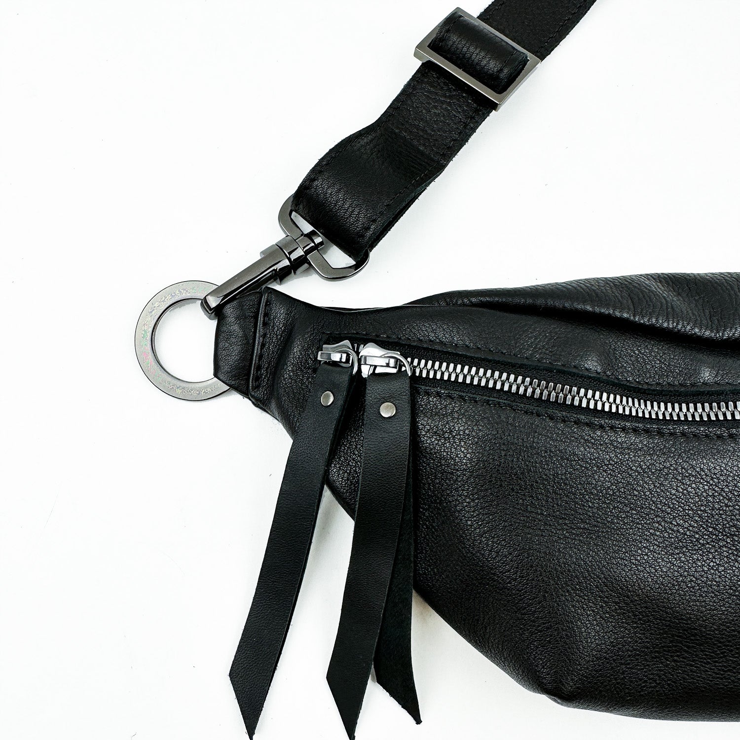 Close-up of the STELLA - BLACK sling bag from evokewomen, showcasing its luxurious black leather with a zipper and two elegant leather pull tabs. The adjustable strap is fastened with a metal ring and buckle, providing a blend of style and functionality.