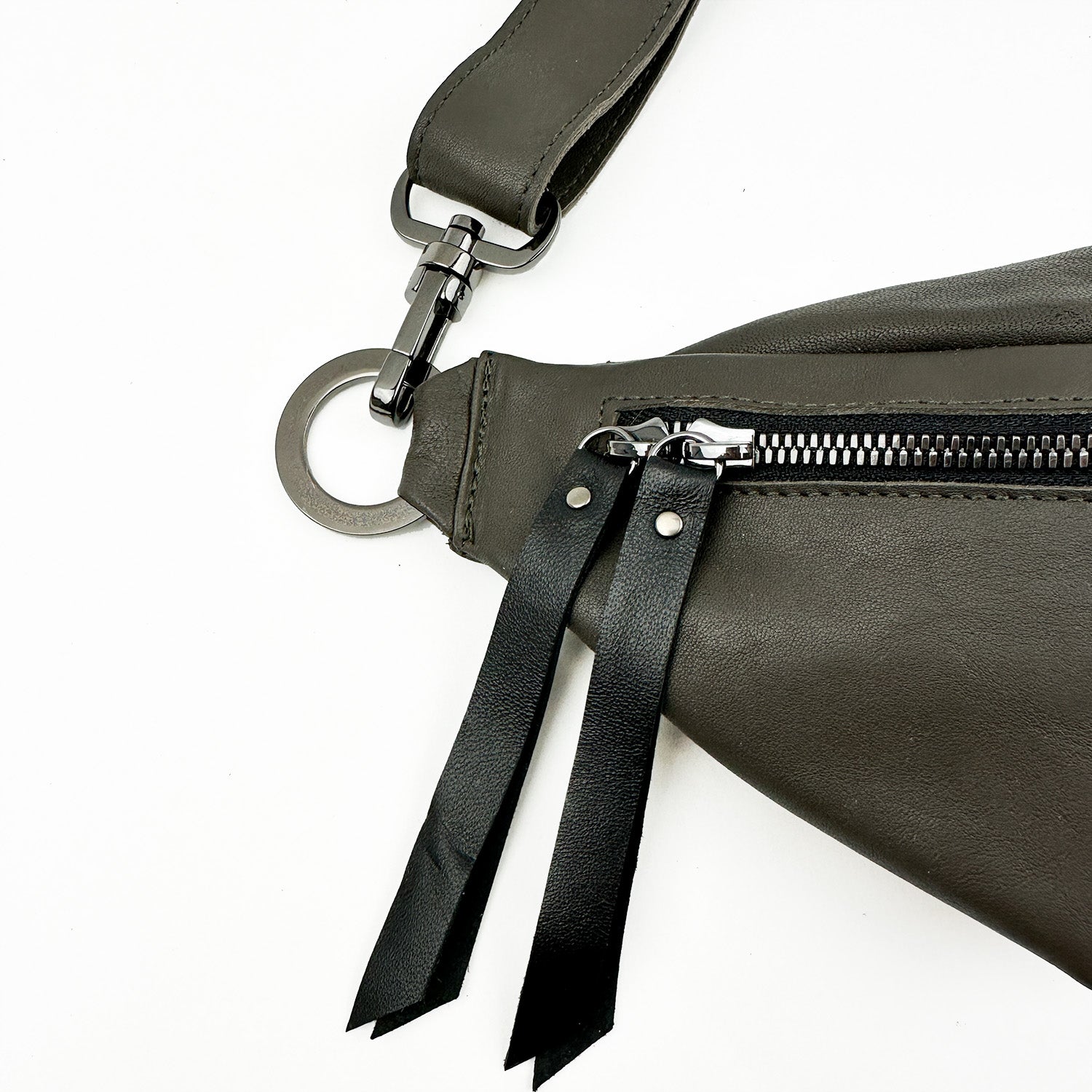 The STELLA - OLIVE-GREY sling bag by evokewomen showcases a close-up of its dark gray leather design, featuring an adjustable strap and metal zipper complemented by two black leather pull tabs. The strap is elegantly secured with a metal ring and clasp.