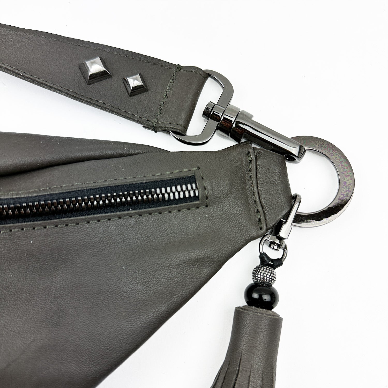 Close-up of a corner of the STELLA - OLIVE-GREY leather sling bag by evokewomen, featuring a zipper, metal studs, buckle, and tassel keychain. The adjustable strap enhances both versatility and style in this chic accessory.