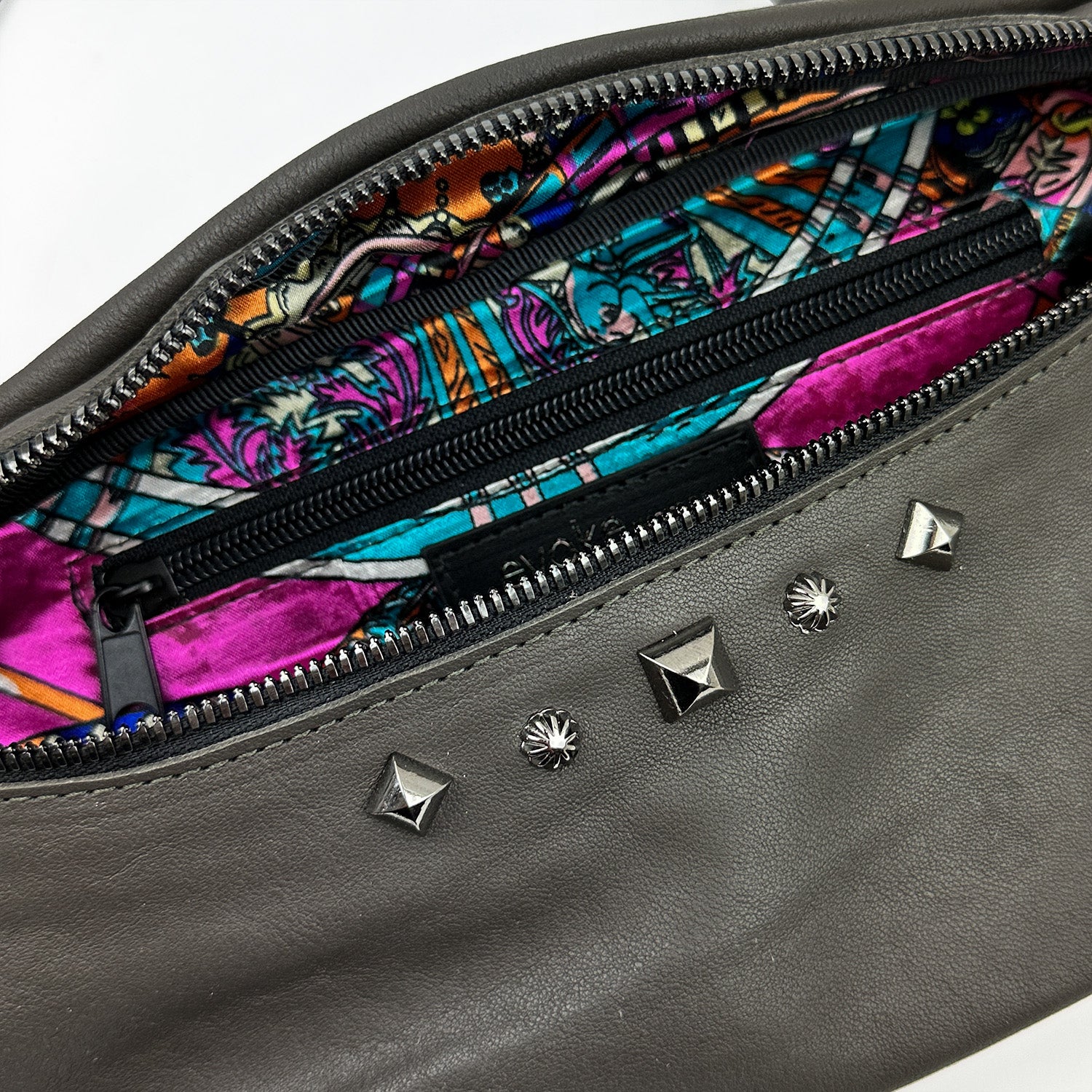 Introducing the STELLA - OLIVE-GREY clutch by evokewomen: crafted from gray leather with striking metal studs, this clutch boasts a vibrant patterned interior lining. It features a convenient zipper pocket and an adjustable strap for versatile styling options.