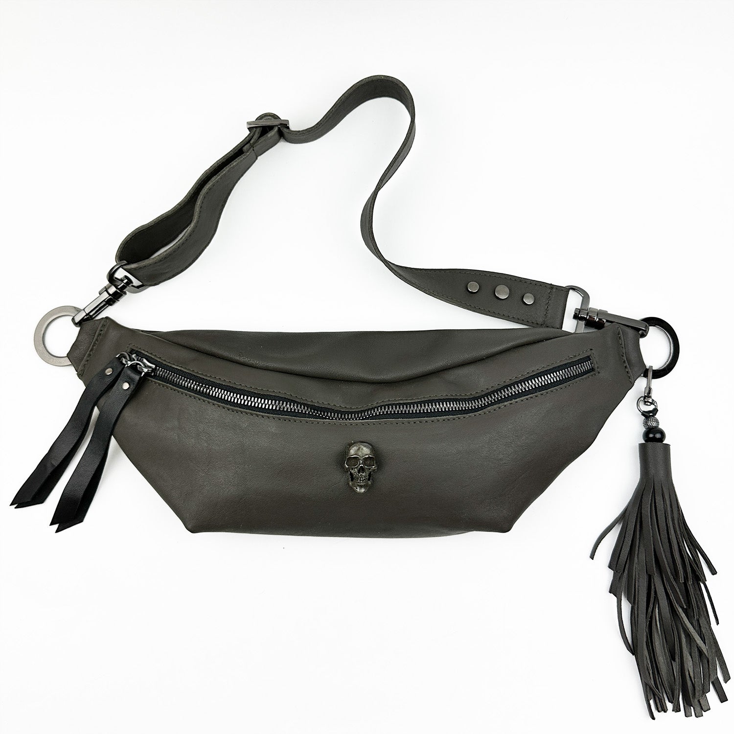 STELLA in olive-grey by evokewomen is a leather waist bag featuring a silver skull emblem, a zipper closure, and a detachable strap adorned with black tassels.