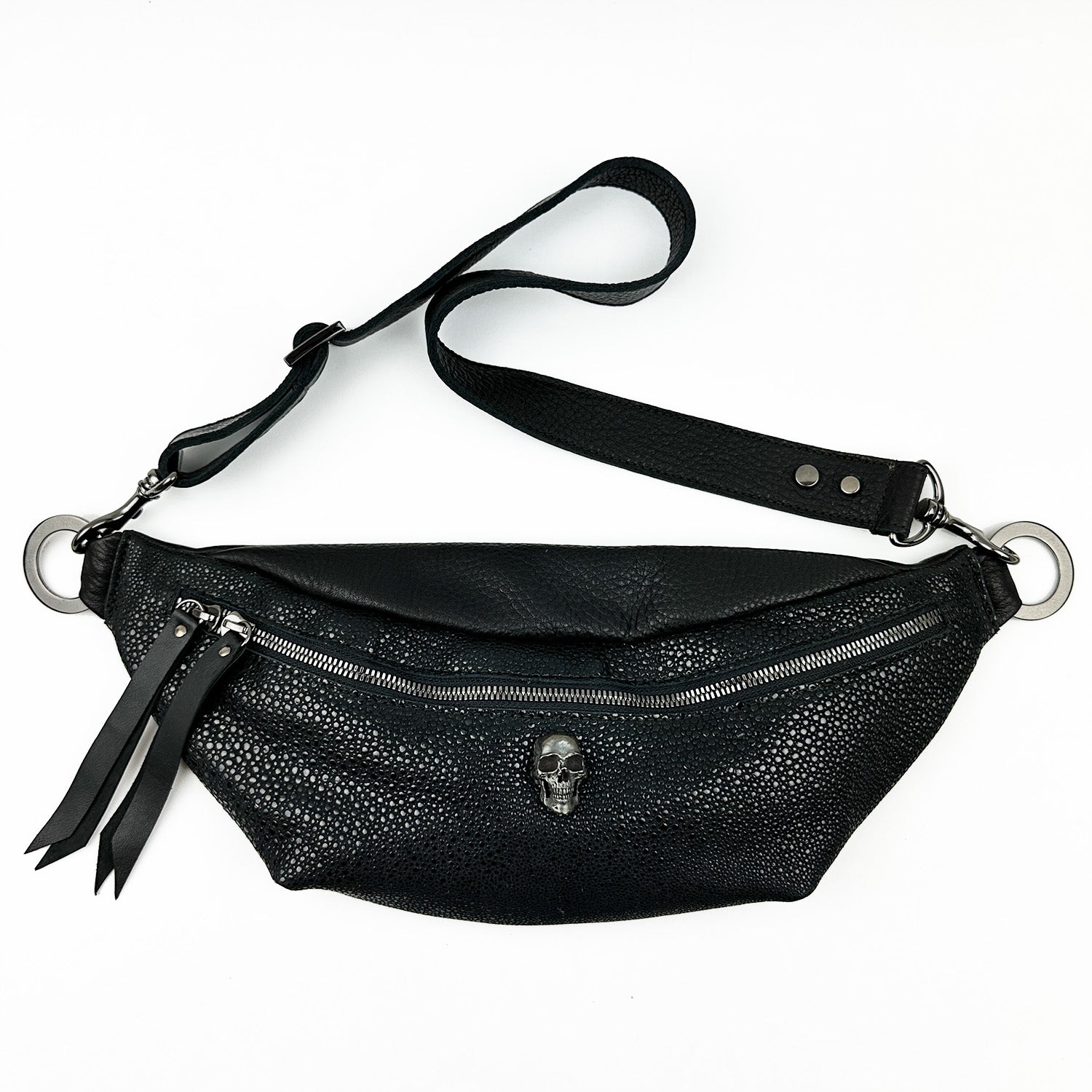 The STELLA - BLACK STINGRAY by evokewomen is a black leather sling bag featuring a single zipper, skull-shaped embellishment, and an adjustable strap.