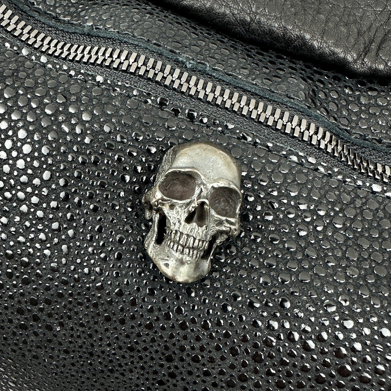Close-up of a metal skull decoration on the textured black leather STELLA - BLACK STINGRAY crossbody bag by evokewomen, featuring a visible silver zipper.