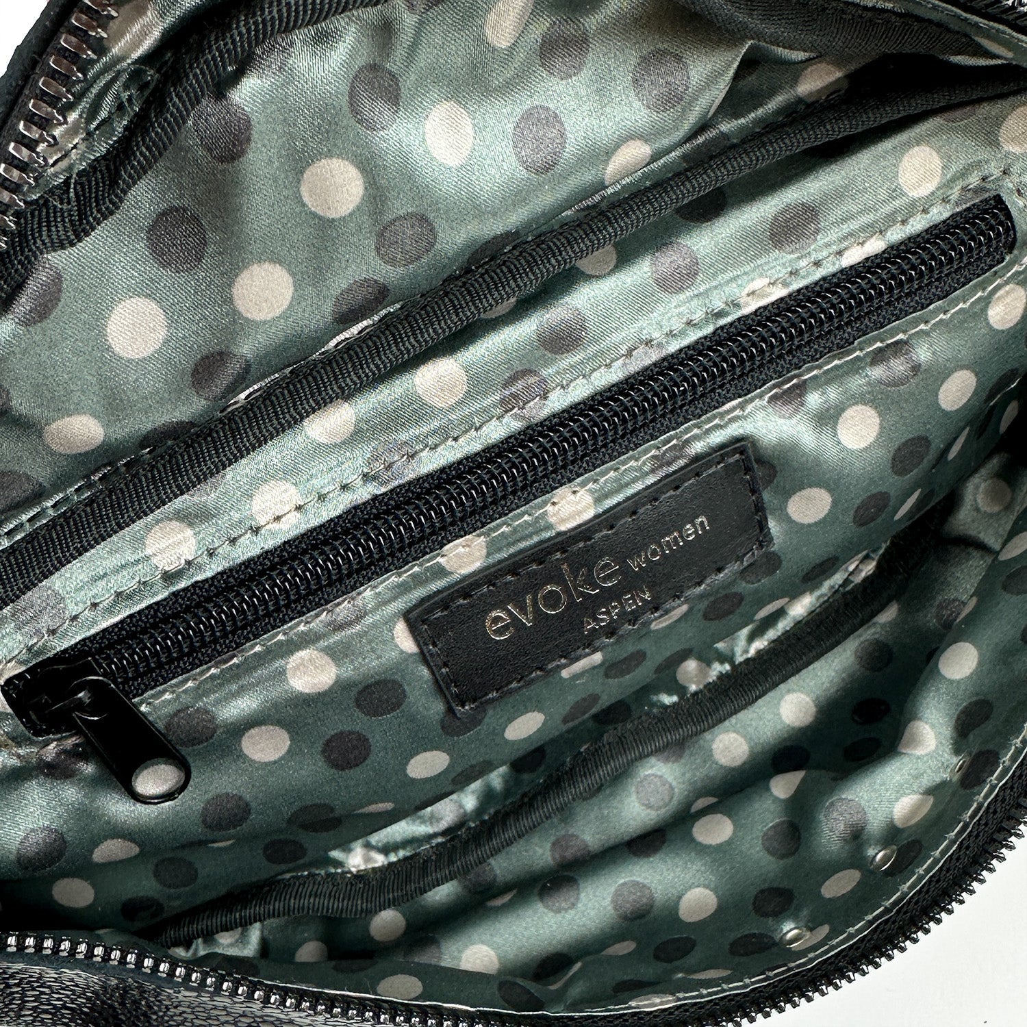 A close-up of an open cross body bag with a polka dot lining, displaying a label that reads "evokewomen STELLA - BLACK STINGRAY" and exposing a zippered interior pocket.