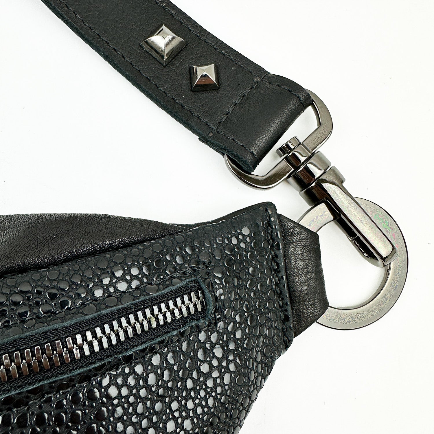 A close-up of the STELLA - BLACK STINGRAY sling bag by evokewomen, showcasing faux stingray leather with a metal zipper, silver clasp, and studded strap against a white background.