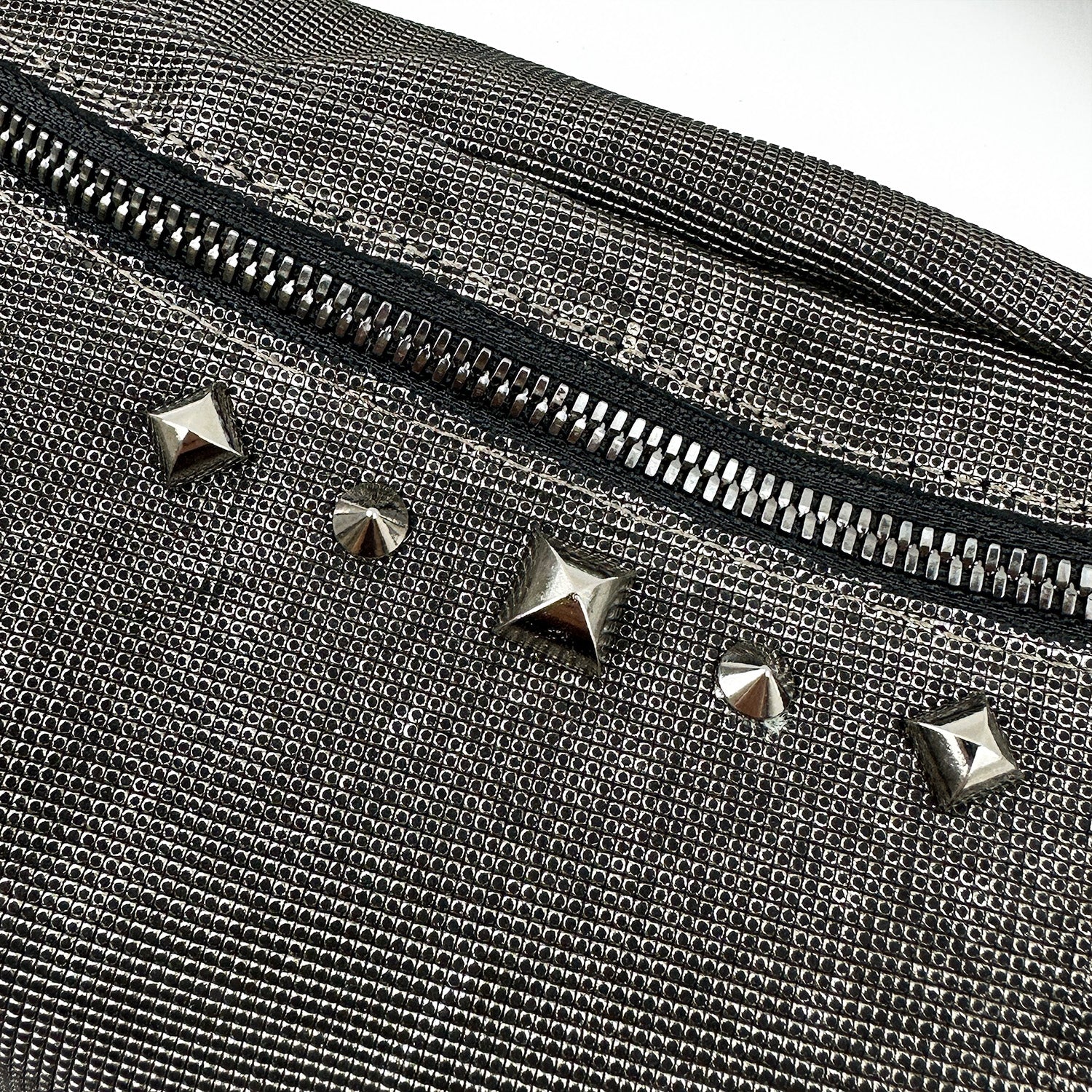 Close-up of the STELLA - GUNMETAL TEXTURE, a stylish cross body bag by evokewomen, showcasing its textured metallic fabric with a visible zipper and five silver studs in various shapes.