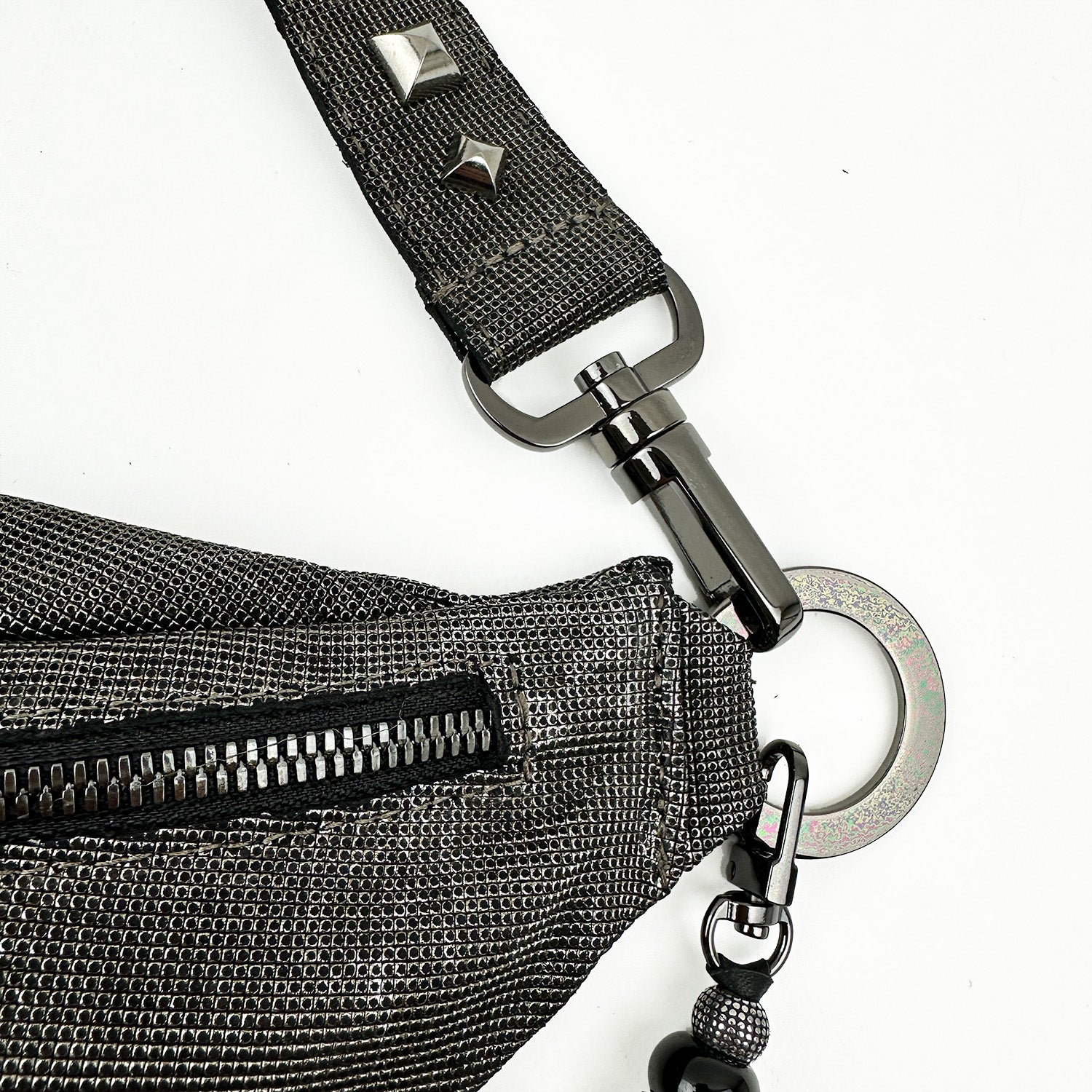 A close-up shot of the STELLA - GUNMETAL TEXTURE crossbody bag by evokewomen, showcasing its metallic textured strap with metal hardware such as a zipper, a hook, and stud details against a white background.