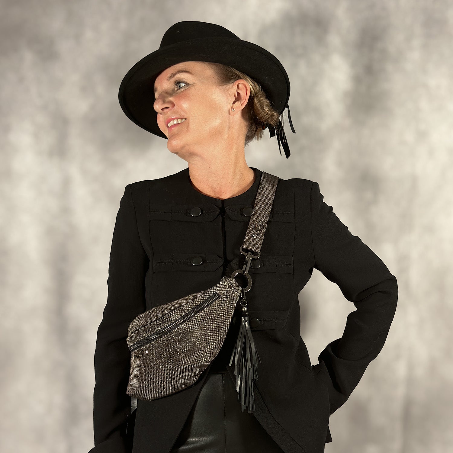 A person sporting the STELLA - GUNMETAL TEXTURE sling bag by evokewomen, wearing a black hat and jacket, stands against a gray backdrop.