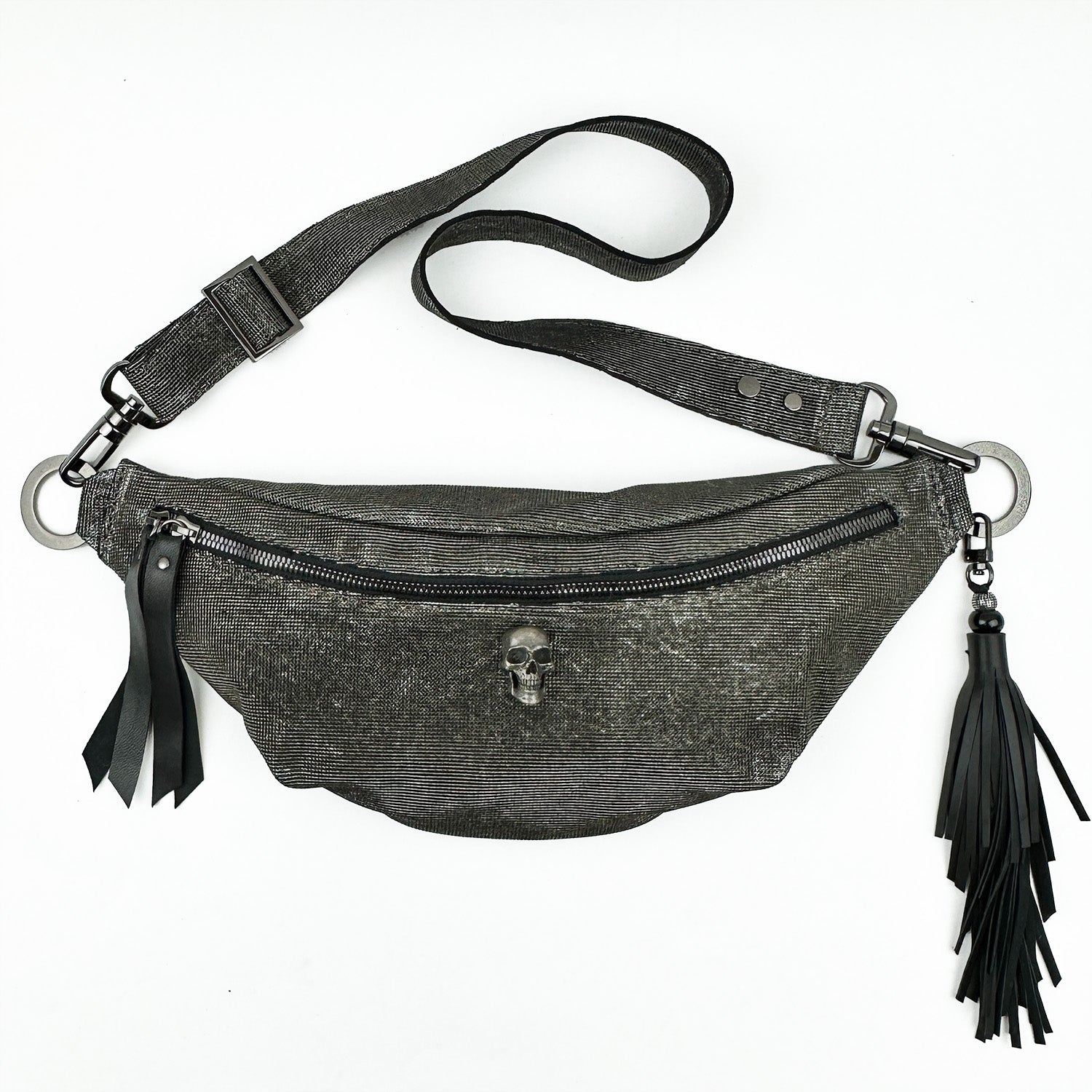 The STELLA - GUNMETAL TEXTURE by evokewomen is a black leather sling bag featuring a zipper, skull embellishment, and tassels on the side.