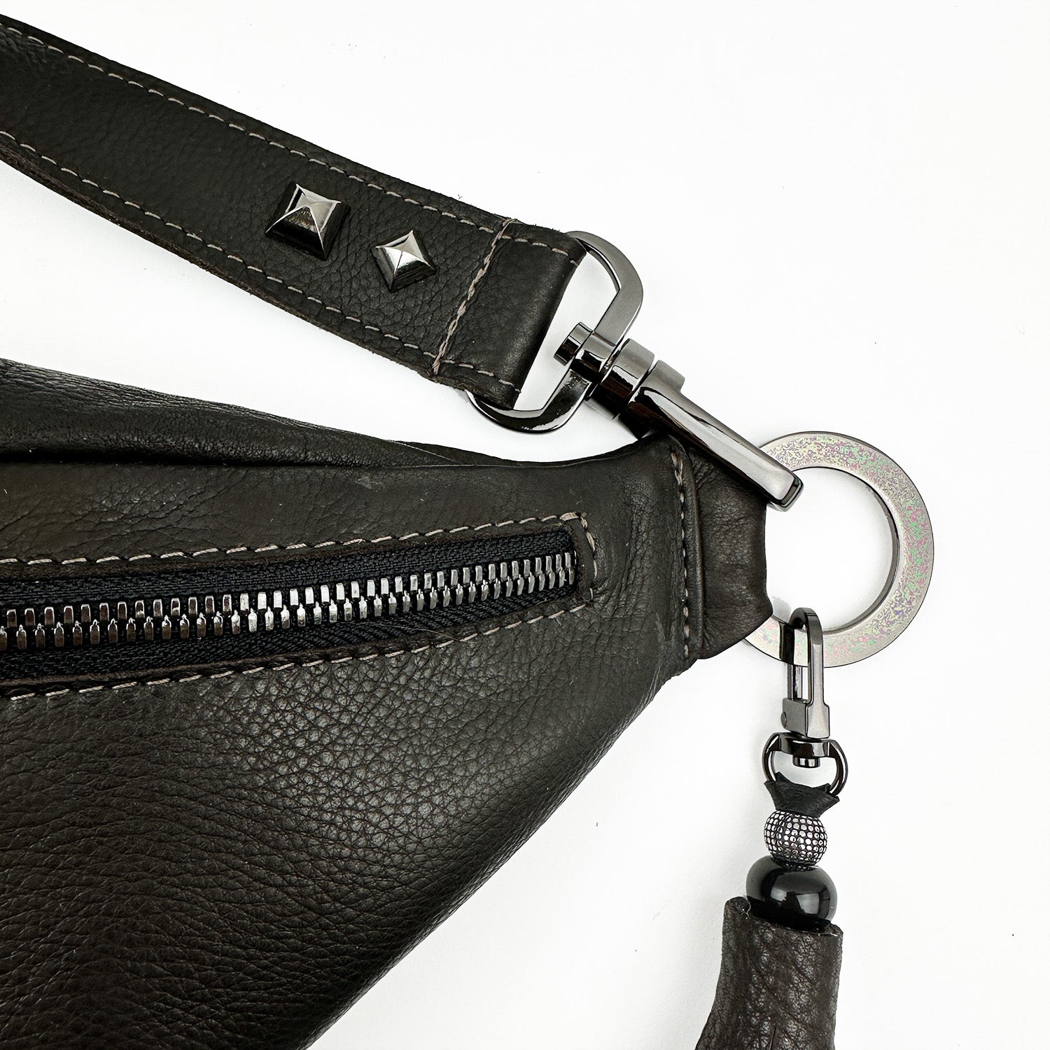 A close-up of the STELLA - BROWN sling bag by evokewomen showcases its sleek design in premium Italian leather, complemented by metal studs and a circular ring attachment. The bag features a prominent zipper, an adjustable strap, and a stylish beaded accessory.