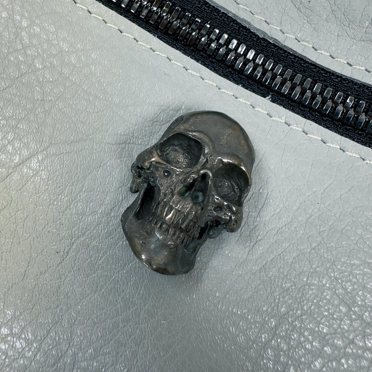 The STELLA - light smoke, a metallic skull sculpture from evokewomen, rests on a textured gray surface near the black zipper of a sleek leather sling bag.