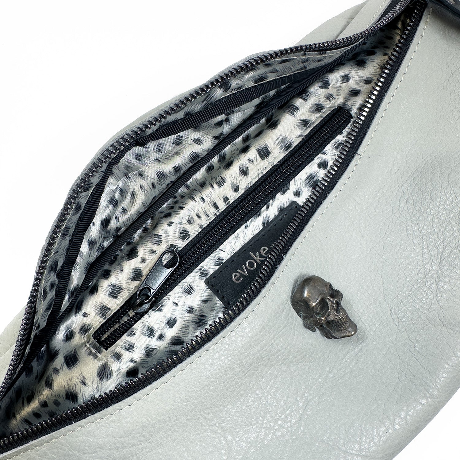 Introducing the STELLA - light smoke from evokewomen: a chic white leather sling bag with a leopard print interior and a visible zipper pocket. Its adjustable strap ensures comfort, while a small skull ornament adds an edgy touch to the exterior.